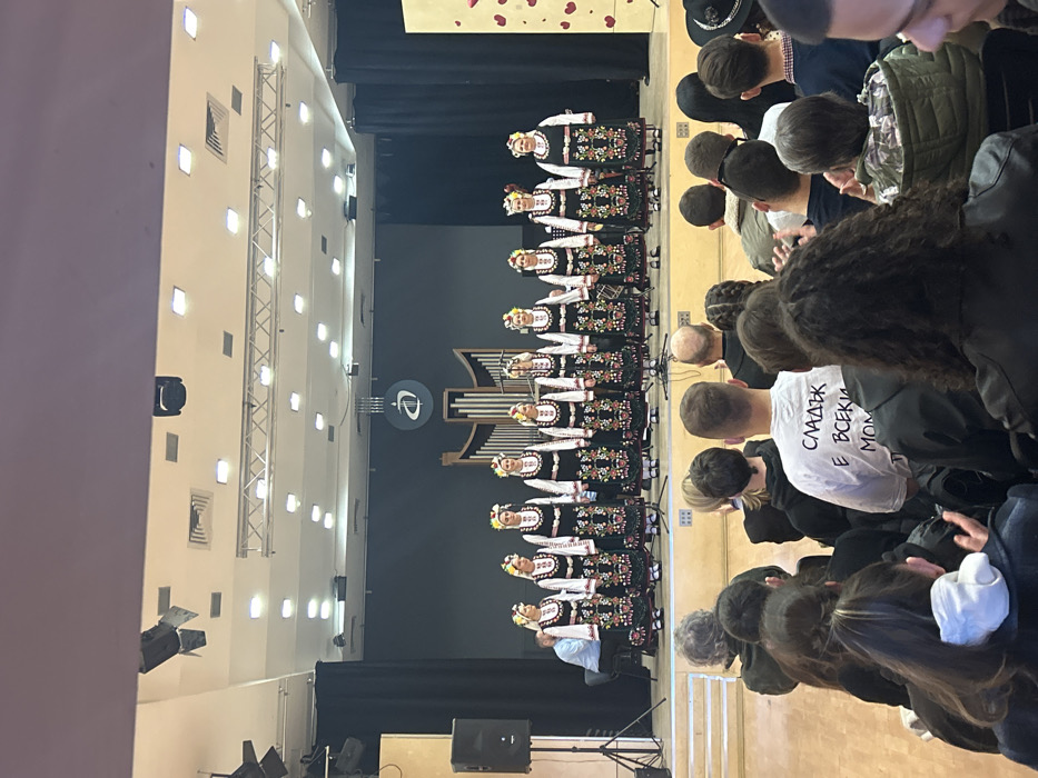 The image depicts an indoor cultural performance in an auditorium, likely within the Plovdiv University "Paisii Hilendarski" in Plovdiv, Bulgaria. This identification is based on the university's stylized 'P' logo displayed on the dark stage backdrop, positioned above an ornate, organ-like structure.
On the elevated stage, ten women stand in a linear formation, facing the audience. Each woman is dressed in traditional Bulgarian folk attire, consisting of a white long-sleeved blouse, a black embroidered vest or apron with colorful floral patterns, and a floral wreath worn on her head. Multiple microphones are positioned in front of the performers. To the stage's left, a male individual in a light blue shirt is seated at a keyboard instrument. A large black speaker on a tripod stand is located left of the stage. To the far right, a light-colored rectangular panel adorned with multiple red heart shapes is visible. Overhead, theatrical lighting fixtures are suspended from a truss system against a light ceiling.
The foreground is occupied by an audience, primarily viewed from behind, seated in rows of chairs and observing the stage. Approximately twenty individuals are clearly visible. One male audience member in the mid-foreground wears a white t-shirt with black Cyrillic text on the back, reading "СЛАДЪК Е ВСЕКИ МОМ." The environment suggests a formal concert or cultural event.