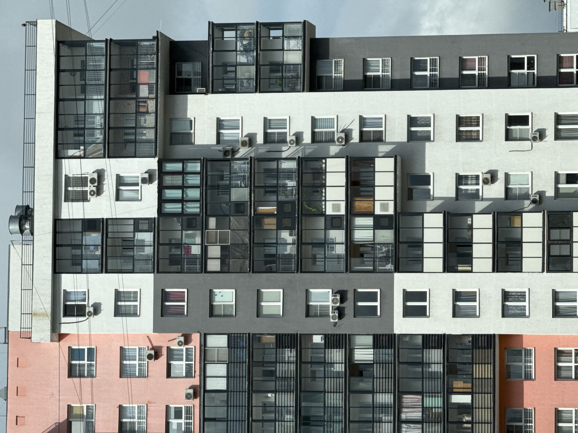 The image shows a modern apartment building in Chelyabinsk, Russia, with a section of the facade painted pink, another section gray, and a central section white. The building has many windows and balconies, some of which are enclosed glass structures. Several air conditioning units are visible on the exterior walls. The sky is cloudy.