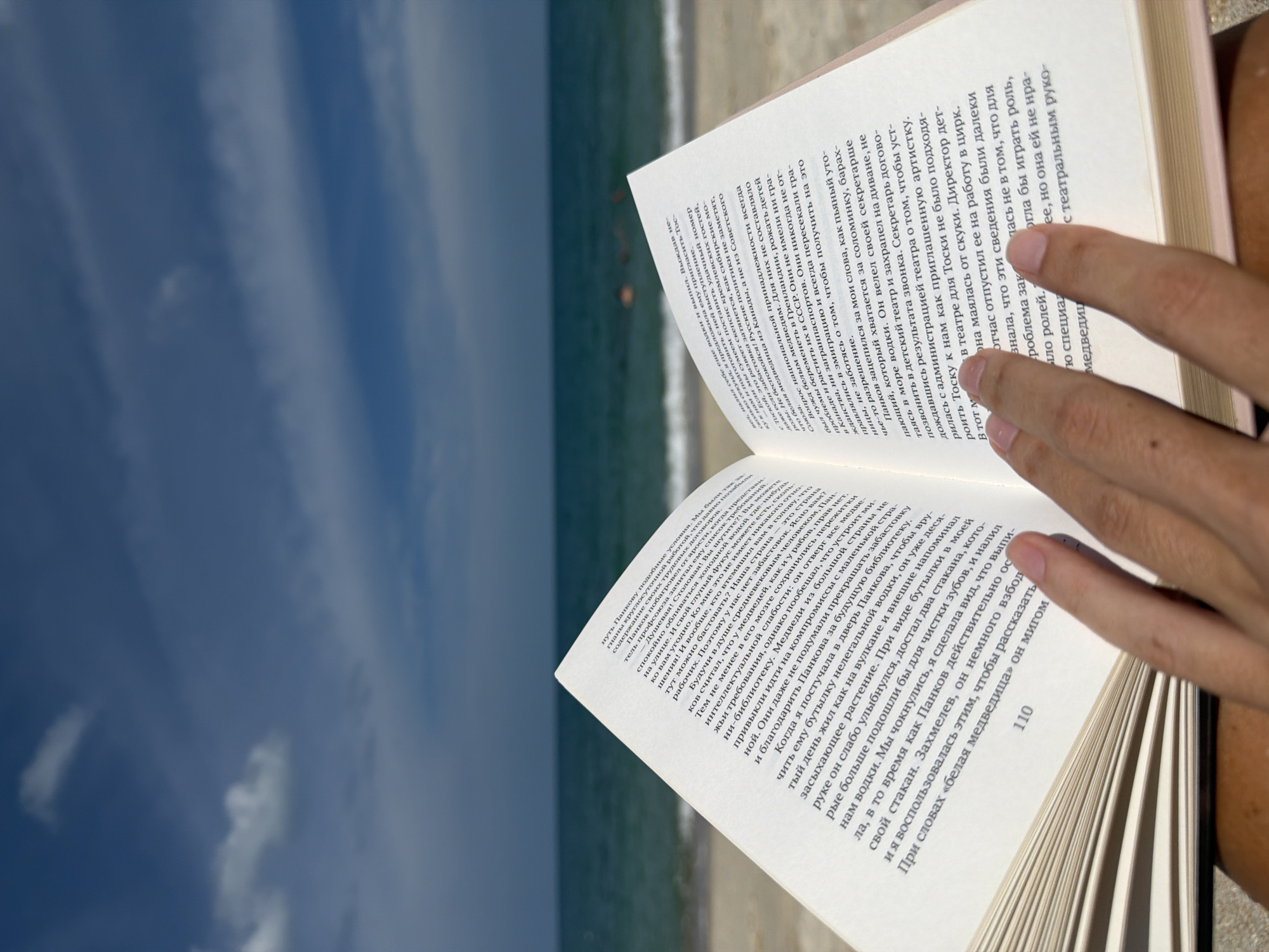An overhead, close-up, eye-level shot captures a person holding an open book on a beach. The book's pages display Russian text, with one page numbered "110." The person's hands are visible, gently holding the book open. In the background, the azure ocean meets a sandy beach, with the faint silhouettes of people swimming in the distance. The scene is bathed in natural light, suggesting an outdoor setting in a tropical location, possibly Ko Samui District, Thailand, given the warm, sunny atmosphere.