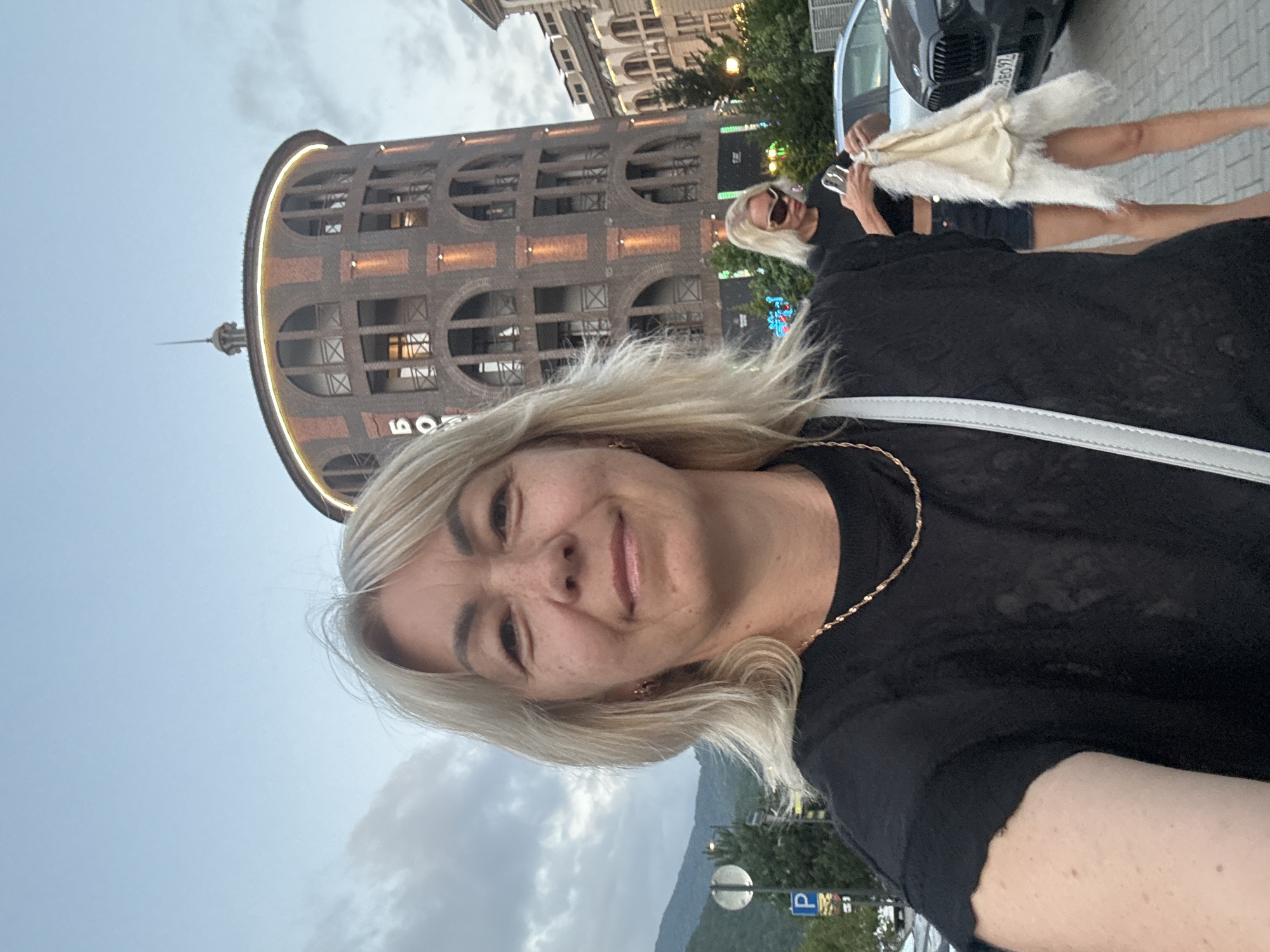 This is a selfie of a blonde woman with light skin and freckles. She is wearing a black t-shirt and a thin gold necklace. Her hair is shoulder-length and blonde. She is smiling and looking at the camera. Behind her, another woman with blonde hair and sunglasses is holding a white furry garment. She is wearing a black top and denim shorts. In the background, there is a distinctive round, brick building with arched windows and what appears to be an inscription on its side. The setting appears to be a street or plaza in Krasnaya Polyana, Russia, with a car visible and a mountainous landscape in the distance under a cloudy sky. The time of day appears to be late afternoon or early evening, with the building's lights illuminated. There is a parking sign visible in the background.