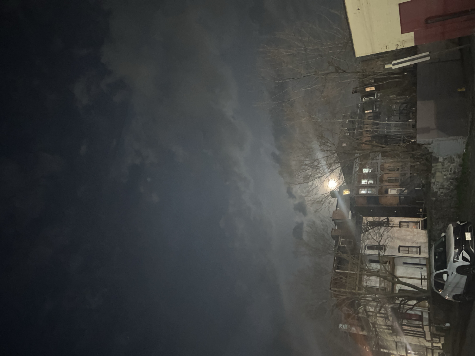 At night, the image shows the City of Newburgh, United States of America, where a full moon illuminates the city skyline. The sky is overcast, with thick clouds obscuring parts of the moon. Buildings of varying heights are visible, silhouetted against the night sky. Bare trees are scattered throughout the scene, their branches stark against the dim light. A white car is parked on the street. Streetlights and illuminated windows cast a soft glow on the surroundings.