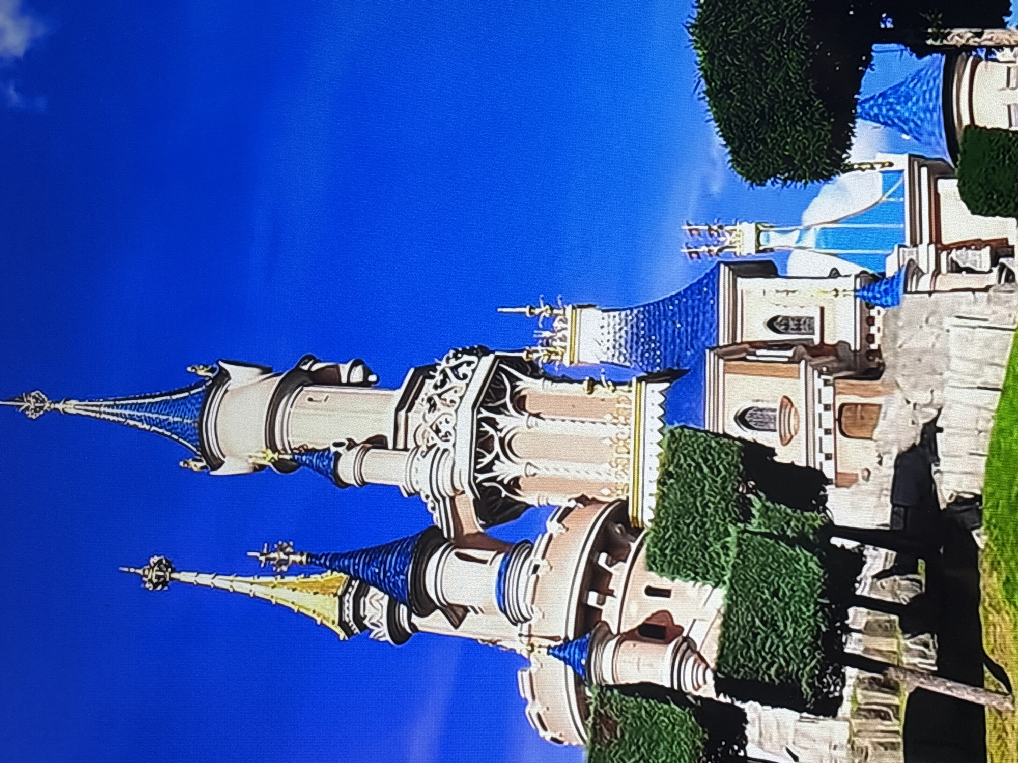 The image depicts Sleeping Beauty's castle, a fairytale castle structure, set against a clear blue sky. The castle features elaborate architectural details, including spires with blue-tiled conical roofs, ornate stonework, and turrets. Green hedges are visible around the base of the castle, and other castle buildings are positioned nearby. The location is Disneyland Park.