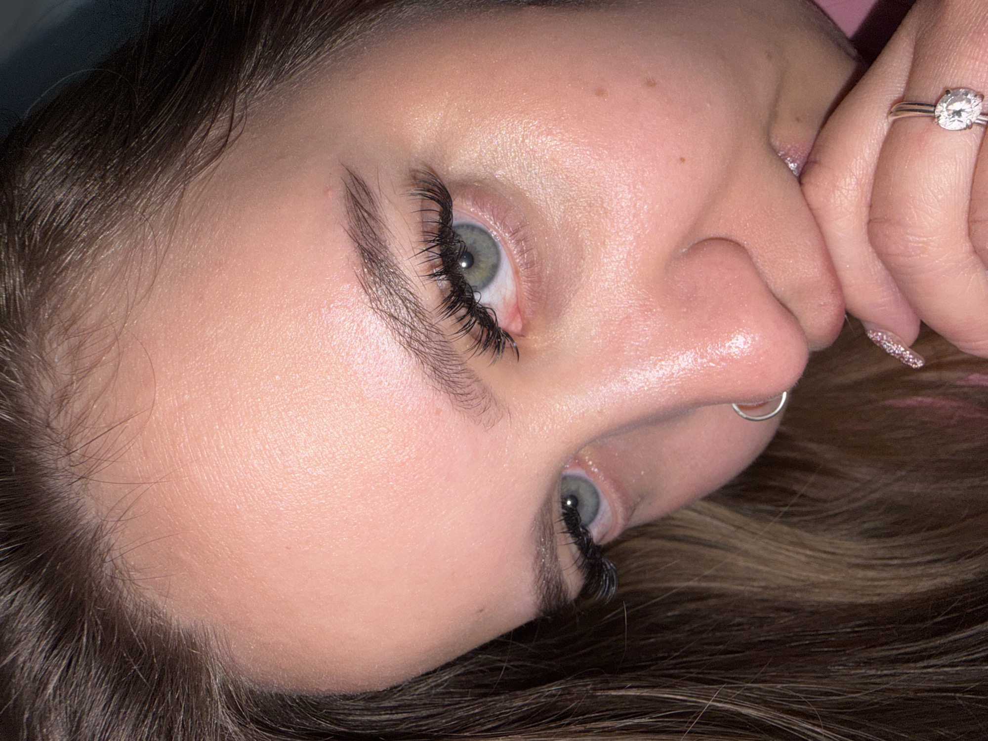 A close-up, angled view of a woman's face. She has fair skin, light brown hair, and striking green eyes enhanced with prominent eyelash extensions. A silver nose ring is visible in her left nostril. Her manicured fingers are partially obscuring her mouth, and a diamond ring is visible on her right ring finger. The background is blurred and suggests a soft, pink surface. There is no indication of Port-Katon, Russia.