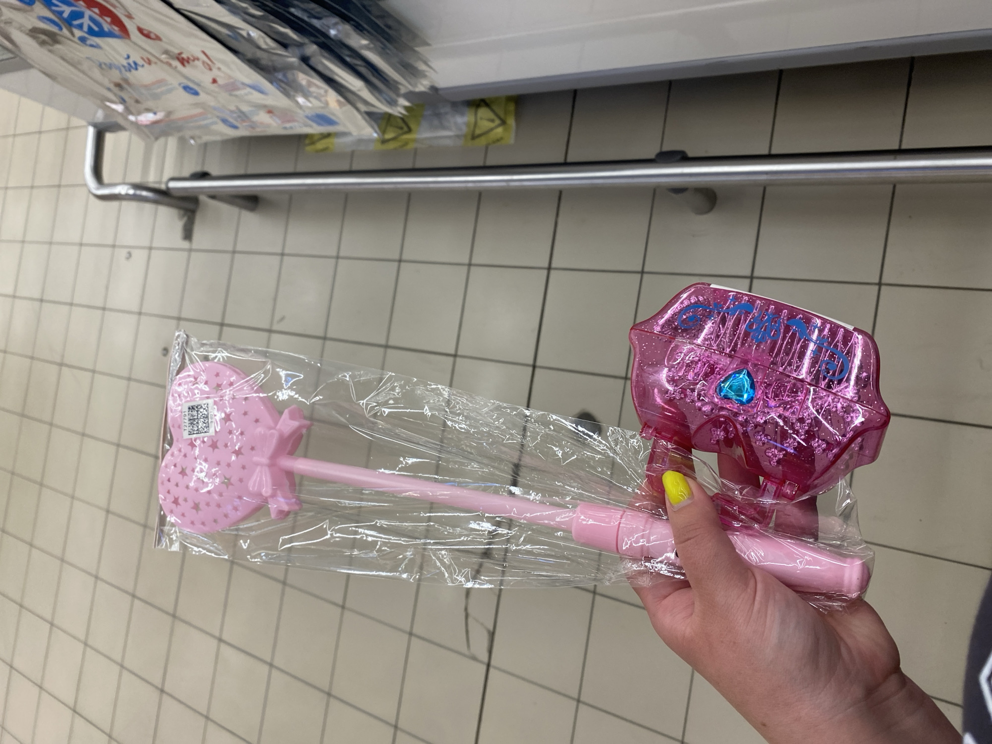 In Kuzmolovskiy, Russia, a person's hand with bright yellow nail polish holds two pink, heart-shaped toys. The larger toy, a wand, is encased in clear plastic packaging and features a pink heart with stars and a bow at the top of a long, pink handle. To the right of the wand is a smaller, pink, heart-shaped case with a blue gem and the word "Crystal" written in blue script. The background consists of a tiled floor and a metal shopping cart.