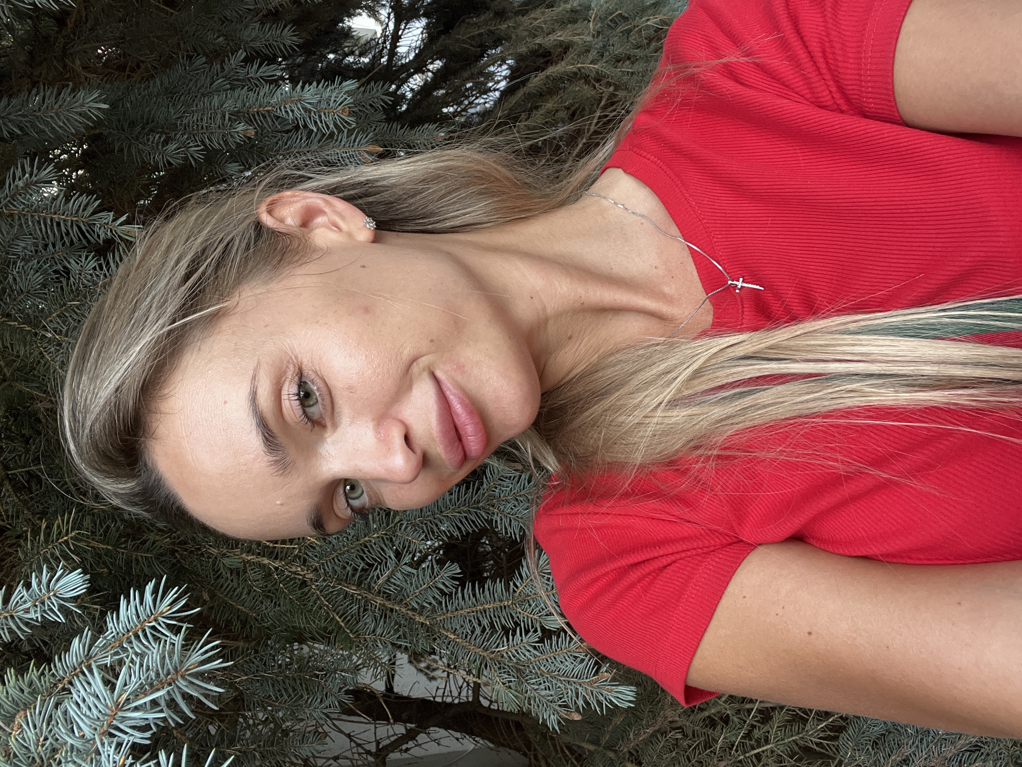 The image is a close-up, waist-up selfie of a woman with blonde hair, green eyes, and light skin. She is wearing a red, ribbed t-shirt and a silver necklace with a cross pendant. She has on small stud earrings. The woman is outdoors, with a backdrop of blue spruce branches. The location of the image is stated to be Fontanka, Ukraine, though this cannot be visually confirmed from the image content alone. The woman is looking towards the camera with a slight smile.