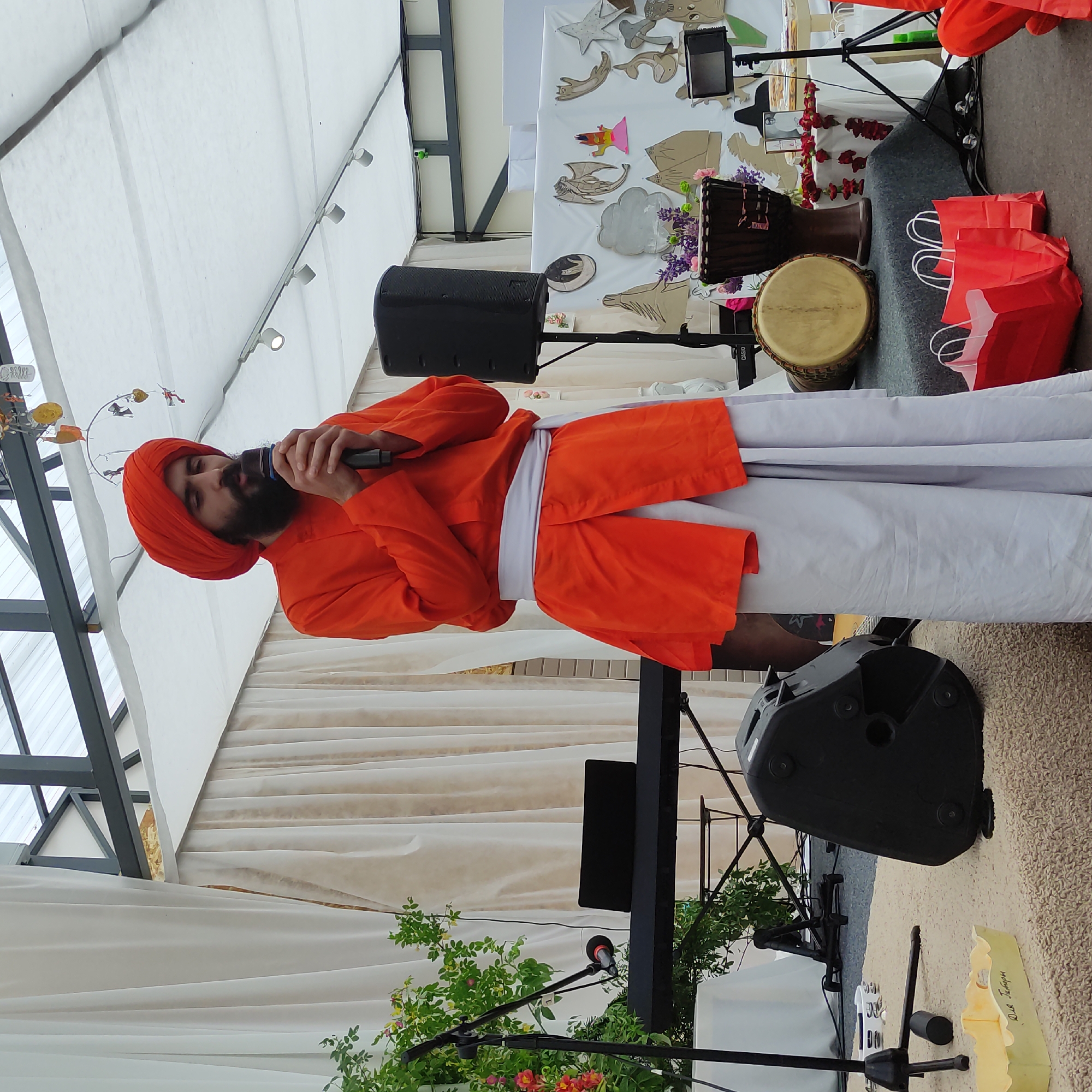 In Zhirnosovo, Russia, a man wearing a bright orange turban and matching tunic stands on a platform, holding a microphone and speaking. He wears a white sash around his waist, and his lower garment is a light gray. He is positioned in front of a stage decorated with white drapes and a white canopy overhead. To his left, a black keyboard is visible, partially obscured by a microphone stand and lush green foliage with pink and orange flowers. To his right, a black speaker column stands tall, and behind him, several drums, including a djembe, are placed on the stage, adorned with purple flowers. Further back, a white cloth is spread with various cutouts of animals and shapes, and a small framed photograph is displayed. The floor of the stage is covered with a gray mat, while the area in front of the stage has a beige, textured rug. Several red paper shopping bags are present on the floor. The scene appears to be an outdoor performance or event.