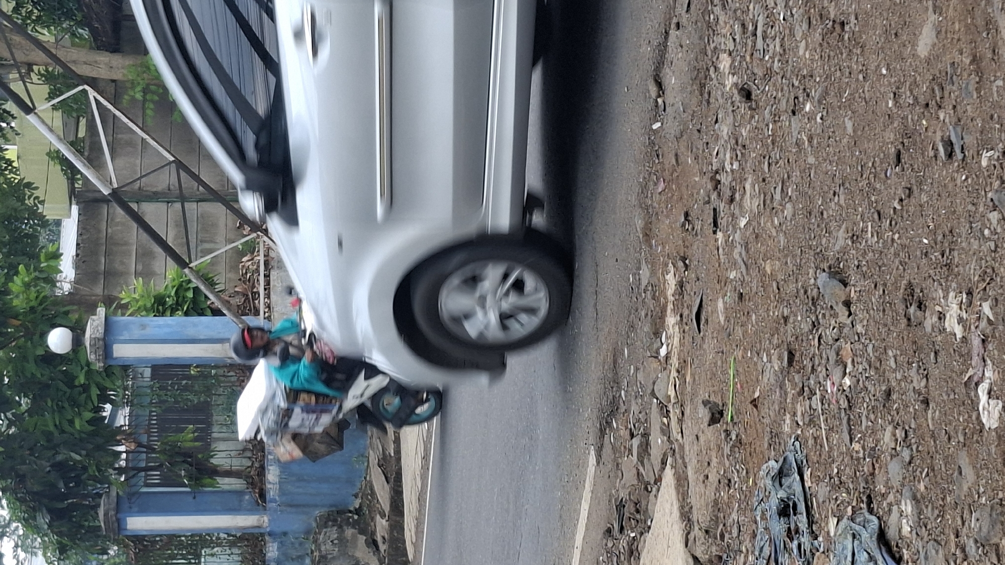 A person wearing a turquoise jacket and a helmet is riding a motorcycle loaded with boxes and other items on a street in Cilaku, Indonesia. The motorcycle is in motion, as indicated by the blur. A silver car is visible in the foreground, also in motion. The background shows a blue and white building with a fence and some trees. The ground on the side of the road is rough and appears to be covered with debris. The scene depicts a typical street in a semi-urban environment, with traffic and everyday activities taking place.