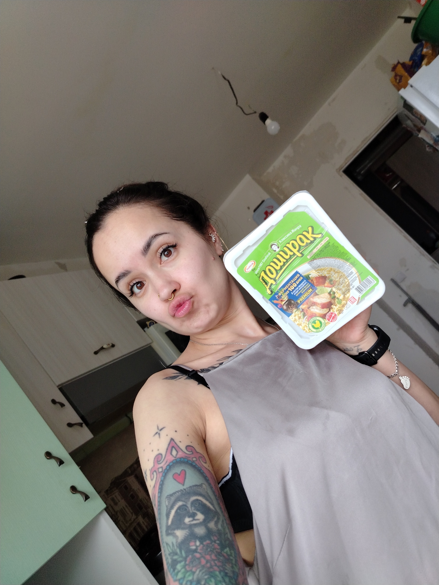 A medium-shot portrait captures a woman indoors, likely in a kitchen setting in Noviy, Russia, holding up a packet of Doshirak instant noodles. The woman, with dark hair tied back, looks directly at the camera, making a playful pouting face. She has prominent eyebrows, long eyelashes, and a golden septum piercing. Her left arm, featuring a colorful sleeve tattoo of a raccoon in a framed mirror with a heart, is visible. She wears a light grey top over a black bra and has a black watch and a bracelet with a foot-shaped charm on her right wrist.
The background shows various elements of a domestic space. To the left, light green and light grey kitchen cabinets are visible. The ceiling above her has exposed wiring and a bare lightbulb hanging, with portions of the wall appearing unfinished or marked.
The packet of instant noodles, prominently displayed, is white with a green label. Visible text on the packaging includes "Doshirak" in both English and Cyrillic script (Доширак), "Since 1987 Doshirak," "Классика Жанра" (Classic Genre), "ФЕРМЕНТАЦИЯ: БОЛЕЕ 100 ДНЕЙ" (Fermentation: Over 100 Days), "PREMIUM," and "Chicken." The packaging also features an image of noodles with chicken.
The image appears to be a selfie or a casual photo, possibly taken during the daytime due to the ambient light, showcasing the woman and her instant meal choice in a somewhat informal domestic environment.