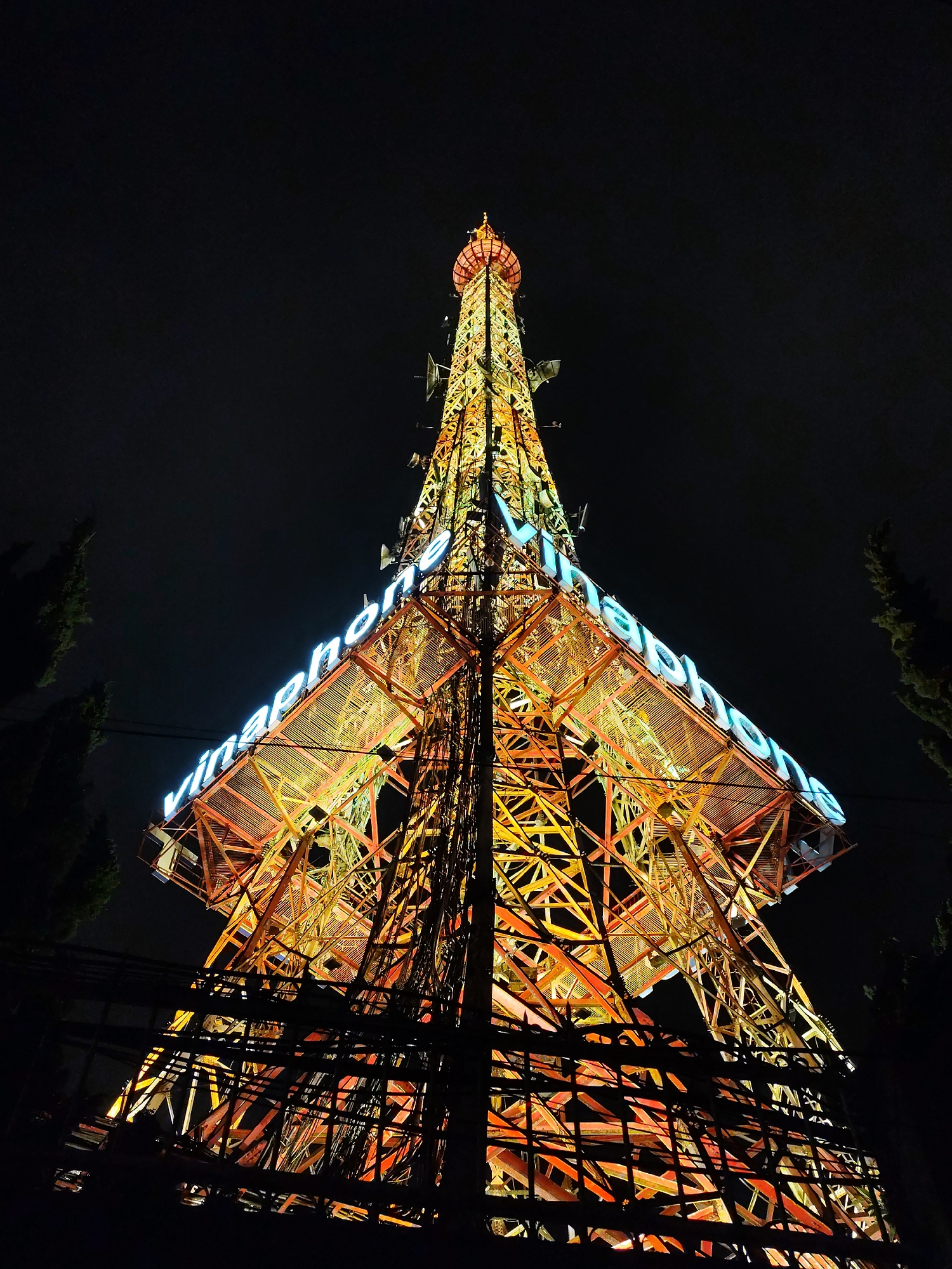 The image shows the VinaPhone tower in Da Lat, Vietnam, at night. The tower is a tall, lattice-structure illuminated with orange and yellow lights. The word "VinaPhone" is displayed prominently in blue neon lettering across the middle section of the tower. It is visible from a low vantage point, with the tower dominating the frame. Electrical wires are visible in the foreground, adding to the complexity of the composition. The background is dark, emphasizing the illuminated tower. Two trees stand on the left and right of the tower.