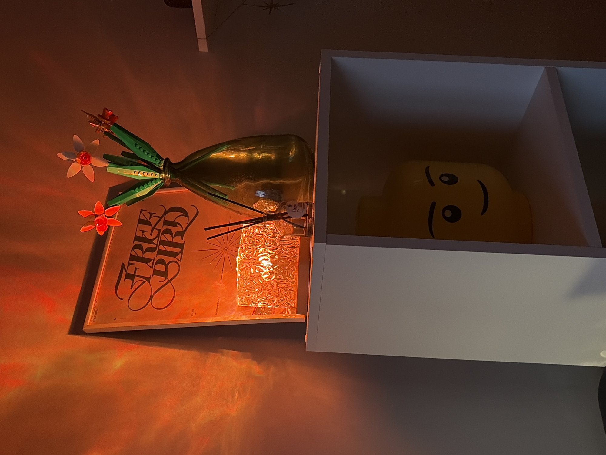 The image shows a still life arrangement with a warm, ambient glow, suggesting it was taken indoors at dusk or night. The main subjects are a bouquet of artificial flowers made from LEGO bricks, a green glass vase, and a vinyl record with the text "FREE BIRD" in stylized lettering. The LEGO flowers, in red and white, are placed in the vase, which appears to be made of recycled glass with a slight green tint and a textured surface.
To the right of the vase, a shelf unit is visible, with the right compartment holding a large, yellow, smiling LEGO head. The shelf unit is white and appears to be mounted on a wall. The background wall is a muted color, with a subtle, star-like pattern visible in the upper left corner.
The lighting in the scene is dramatic, with orange and red hues casting long shadows and highlighting the textures of the objects, especially the glass vase and the record cover. The overall mood is cozy and perhaps a bit retro, amplified by the presence of the vinyl record. There is no indication of weather conditions or specific outdoor location cues, other than the general interior setting. The LEGO head, with its simple, iconic face, adds a playful and whimsical element to the composition. The text "FREE BIRD" on the record cover is the most prominent legible text in the image. The setting appears to be a domestic space, possibly a living room or study.