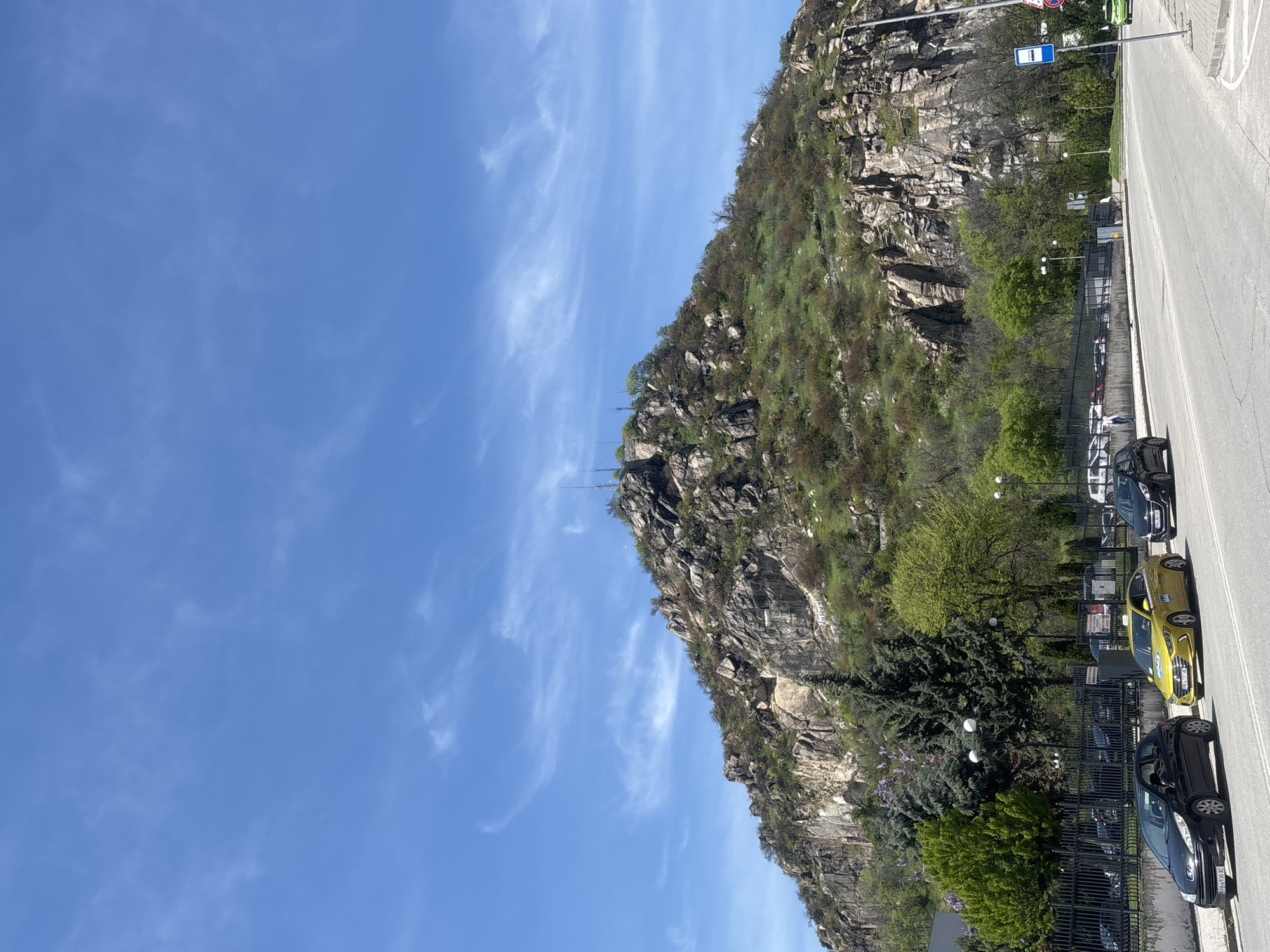 The image, correctly viewed in an upright orientation, depicts a bright daytime scene in Plovdiv, Bulgaria, featuring a prominent rocky mountain adjacent to a road. The upper and left portions of the frame are dominated by a clear blue sky, dotted with scattered, wispy white clouds.
A large, rugged mountain occupies the central and right areas, its slopes composed of grayish-brown rock formations and covered with patches of green vegetation and trees. Near the mountain's crest, several slender structures, possibly antennas or flagpoles, can be seen against the sky.
Parallel to the mountain's base, a paved road stretches across the lower section of the image. Along this road, multiple cars are parked, including a dark-colored sedan in the foreground and a distinct bright yellow compact car. A black metal fence separates the road from the lower mountain slope, which is densely forested with trees and shrubs. Streetlights are visible among the foliage. A blue rectangular sign with a white bus symbol, denoting a bus stop, stands beside the road, alongside other smaller, unreadable traffic signs. The scene is well-lit by sunlight, indicating a clear weather day.