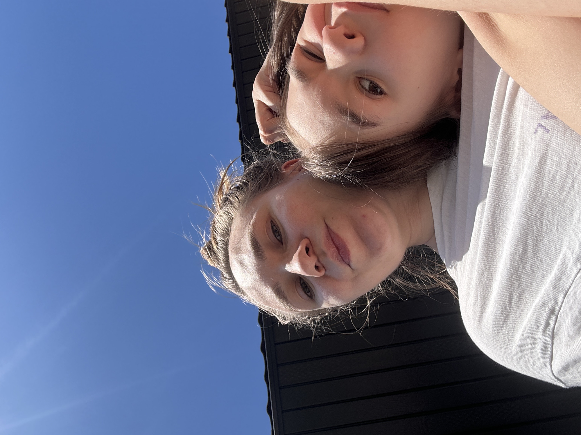 A low-angle, upside-down portrait captures two individuals, likely young women, against a bright, clear blue sky and the dark facade of a building. The image appears to be a selfie or a casual photo taken while looking upwards.
The subject in the foreground has light brown hair, pulled back, and is wearing a light-colored t-shirt. Their face is illuminated by direct sunlight, casting shadows that highlight facial features. They look towards the camera with a neutral expression, showing a slight, gentle smile.
Behind and slightly above the first individual, the second subject has darker brown hair. Their face is also illuminated, showing a similar neutral to slightly smiling expression. They are wearing a light-colored top.
The background consists predominantly of an expansive, cloudless blue sky, indicating fair weather and bright daylight. A dark, possibly black or very dark gray, horizontally planked structure, resembling a building's siding or roof, is visible behind the subjects on the left side of the frame. No discernible text is visible. The scene depicts an outdoor moment, potentially in Kharnavy, Russia, as per the provided context, under a clear sky.