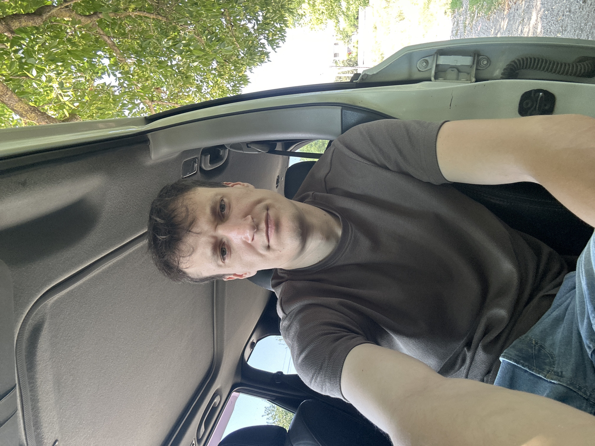 In Makiv, Ukraine, a man is seen from the chest up, sitting inside a vehicle with the passenger door open, facing the camera. He is wearing a brown t-shirt and blue jeans. The interior of the car, including the sunroof, headliner, and seats, is visible. Outside the car, lush green foliage from a tree hangs overhead, and a rural landscape with a dirt road is visible in the background. The lighting suggests it is daytime.