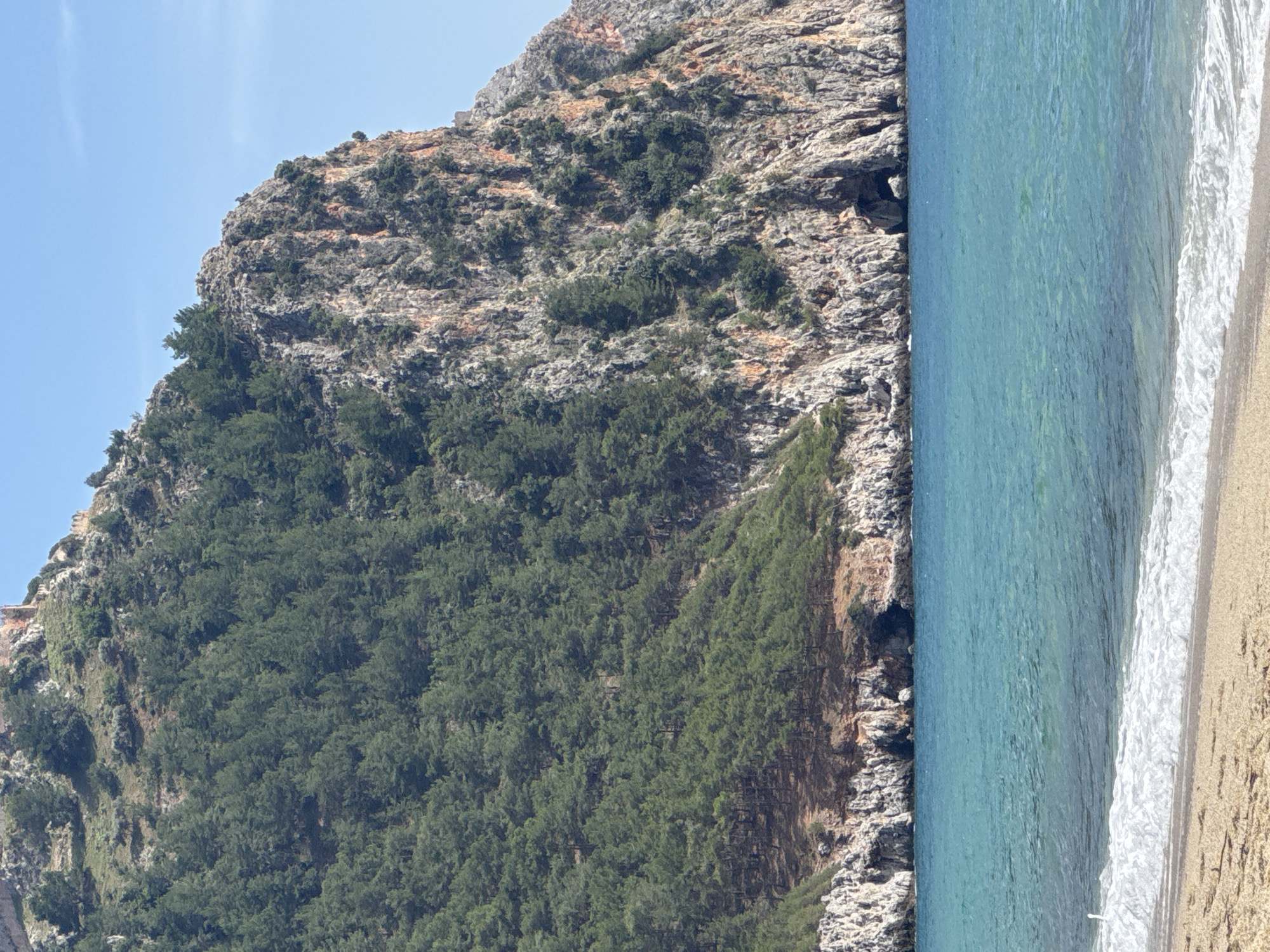 The image is a landscape photo taken in Alanya, Turkey. It shows a rocky mountain covered in trees on one side and a sandy beach and turquoise sea on the other. The sky is clear and blue. The mountain appears to be covered in trees and vegetation, with some rocky cliffs visible. The beach is a strip of sand, with waves crashing on the shore. The water is clear and blue.