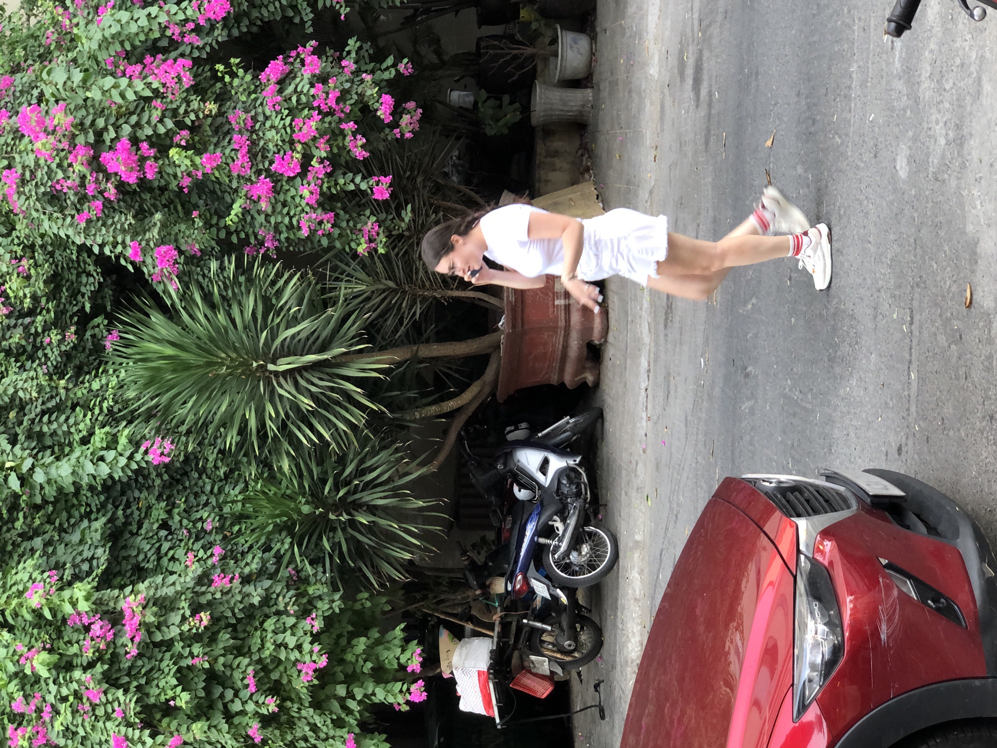 In a city street scene in Da Nang, Vietnam, a young woman in a white t-shirt and white skirt walks while talking on her mobile phone. She is wearing white sneakers with red and white striped socks. In the background, several motorcycles are parked under a canopy of lush green foliage with vibrant pink bougainvillea flowers. A red car is partially visible in the foreground.