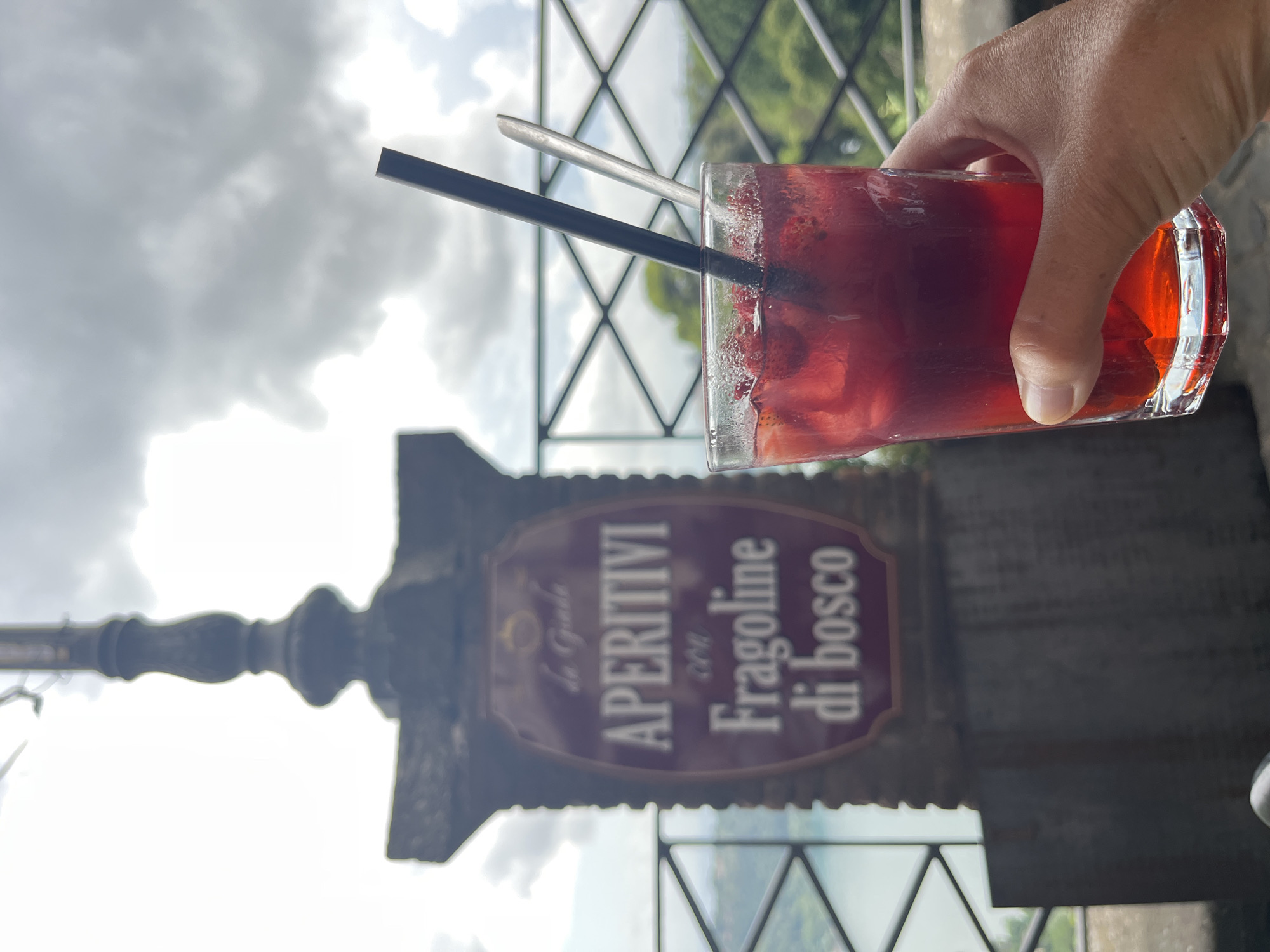 A hand holds a glass of red liquid with ice and strawberries, topped with a black straw and a silver spoon, in the foreground. In the background, a sign with Italian text reads "APERITIVI con Fragoline di bosco" which translates to "Aperitifs with wild strawberries." The sign is mounted on a stone structure, possibly part of a building or landmark. Beyond the sign, a body of water and lush greenery are visible, suggesting a scenic location. The sky above is overcast with dramatic clouds. The scene appears to be in Nemi, Italy, known for its picturesque lake and volcanic crater.