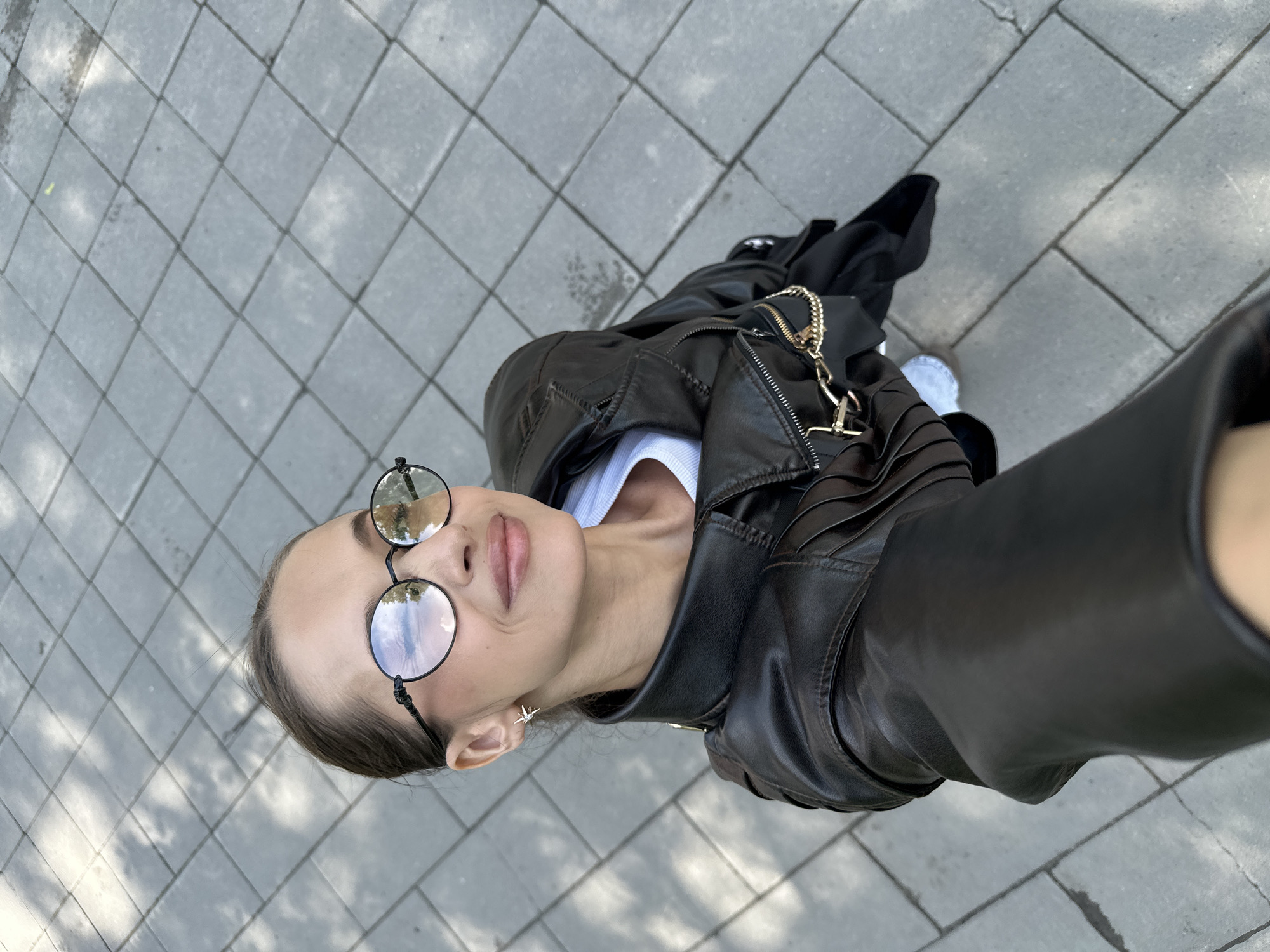 A woman wearing a black leather jacket, a white t-shirt, and round glasses is standing on a tiled sidewalk. She is holding a phone to take a selfie. The sidewalk is made of grey rectangular tiles. There are shadows on the sidewalk from trees. The background is blurred.