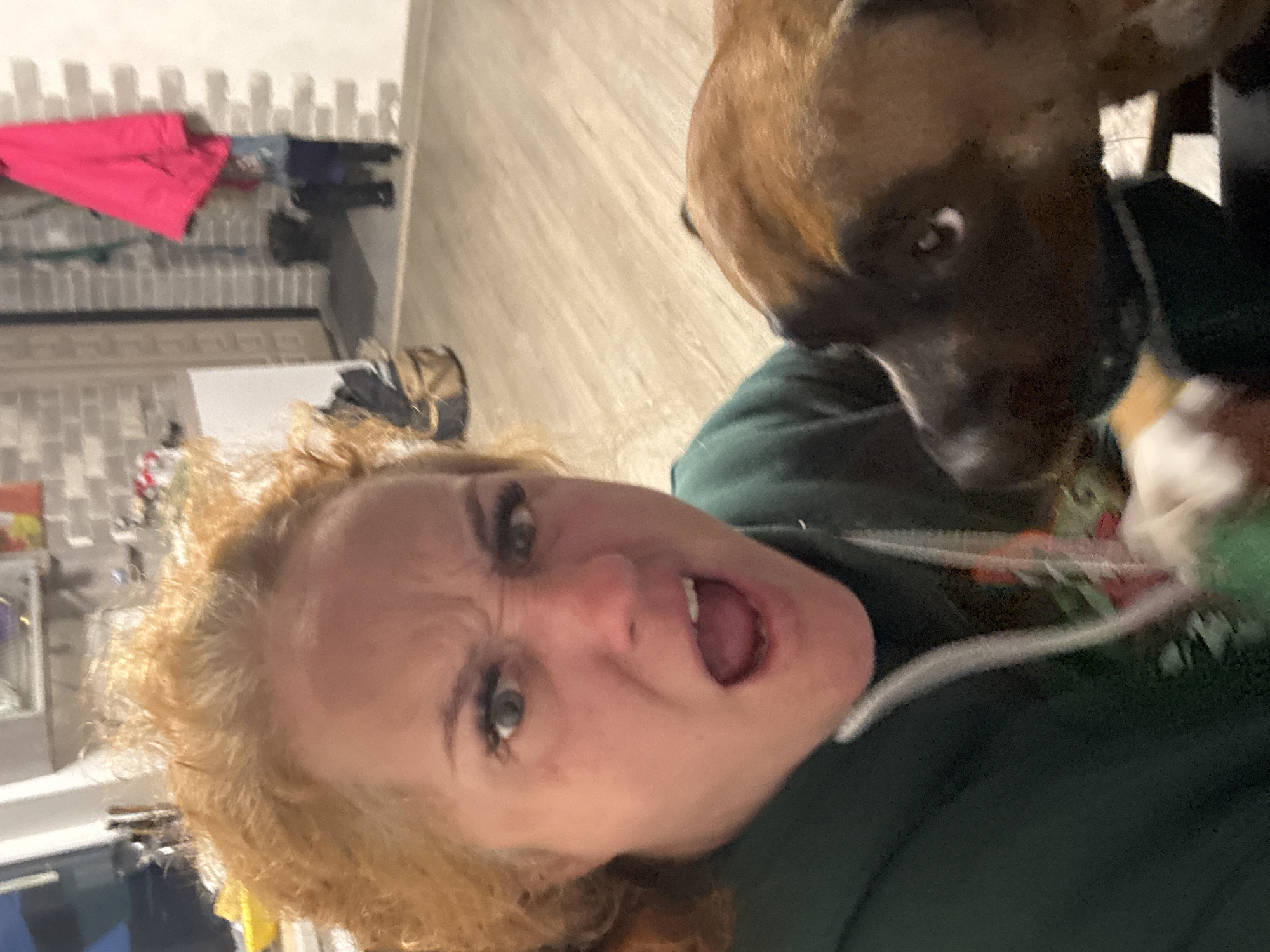 A selfie-style image shows a Caucasian woman with curly red hair and a brown Boxer dog. The woman is making a face with furrowed brows and an open mouth. She wears a dark green shirt or jacket. The dog is positioned to the right of the woman, with its head near her shoulder. The background includes a wall with decorative paneling and a glimpse of clothing hanging. The location is not definitively established as Chaikovsky, Russia, within the frame.