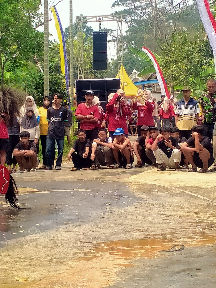 The image captures an outdoor community event in Tamansari, Indonesia, featuring approximately two dozen individuals gathered as spectators. In the immediate foreground, ten young males are squatting in a line on a wet, paved surface, facing left, observing an unseen subject. Behind them, a mixed group of standing adults, including men and women, also observes the activity. Two women wearing red shirts and hijabs are actively holding up smartphones, seemingly recording or photographing. Partially visible on the far left is a person in a performance costume, including a dark, shaggy headpiece and a red, decorated tail-like object. In the mid-ground, a temporary sound system is erected, featuring a metal truss supporting two black line array speakers, with two additional large black speakers on the ground. Two yellow canopy tents and a partial blue structure are visible in the background. Vertical banners, one yellow and blue, and another red and white (Indonesian flag colors), are attached to poles. The overall background consists of dense green trees and foliage. The paved ground exhibits multiple wet patches and scattered light-colored debris.