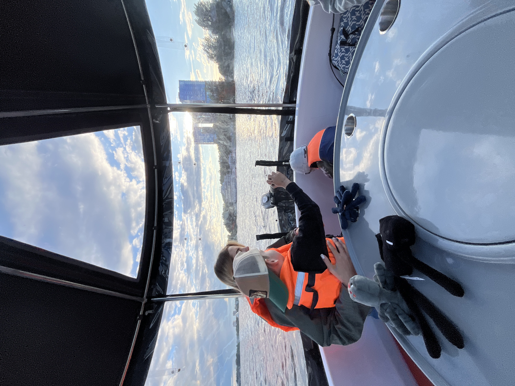 The image depicts a tranquil scene from inside a boat on a body of water in Kazan, Russia, during a late afternoon or sunset. The perspective is from within the boat's cabin or cockpit, looking out towards the cityscape across the water.
Two individuals are seated inside the boat, both wearing bright orange life vests. One person, donning a white and green baseball cap, is positioned towards the boat's controls, with their hands appearing to operate a lever or steering mechanism. Another person is partially visible next to them. The boat's interior includes a light-colored, circular table with cup holders, which beautifully reflects the sky and clouds. Resting on the boat's side are two plush toys: one black, resembling an Enderman character, and another grey, resembling a spider, both from the game Minecraft. A blue and white patterned bag is also visible in the boat's background.
Outside the boat, a wide expanse of water shimmers with reflections of the setting sun and the sky. The sunlight creates a warm glow across the water's surface. On the distant bank, trees and several modern buildings are visible, indicating the urban landscape of Kazan. The sky above is clear with scattered white clouds against a blue backdrop, typical of pleasant weather conditions. The overall impression is one of a peaceful and enjoyable leisure activity, likely a sightseeing tour or a relaxing boat ride on one of Kazan's rivers or waterways. No discernible text is visible in the image.
