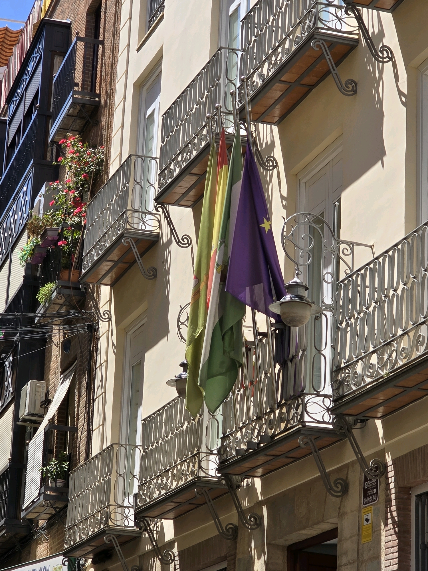 The image shows the facade of a building with multiple balconies on a sunny day. The building has a light-colored stucco exterior and red brick elements. Several balconies feature ornate metal railings and decorative awnings. Colorful flags are hanging from one of the balconies, partially obscuring the view of the building's windows. There are potted plants and flowers on some of the balconies, adding greenery and color to the scene. An outdoor lamp is attached to the railing of a balcony. The lower part of the building has an arched entryway. The presence of Spanish flags and the architectural style suggest the location is in Spain. Given the flags visible, which appear to be the flag of Andalusia and possibly a provincial flag or city flag, it is likely in the city of Jaén, Spain. There are no people visible in the image. The scene is a street-level view of the building's exterior.