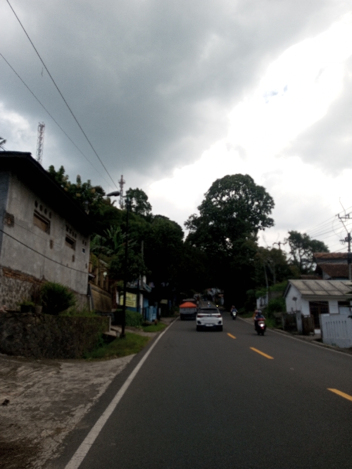 The image captures a street scene in Tagtse Tashidingkha, Bhutan, under an overcast sky. A two-lane asphalt road, marked with a solid white fog line on the left and double broken yellow center lines, ascends and gently curves to the left, extending into the distance. A white SUV and a preceding vehicle with an orange roof structure are traveling away from the viewer. Two motorcycles are also present, one trailing the SUV and another further ahead.
On the left side of the road, a two-story building with light grey walls and an exposed brick and stone foundation stands elevated on a retaining wall. Behind this structure, a densely vegetated hillside rises, featuring a prominent reddish-brown lattice communications tower and multiple utility poles with overhead power lines.
On the right side of the road, a smaller white building with a grey tiled roof is visible in the foreground, with other structures partially obscured by a line of large, dark-leafed trees that border the roadside. The sky is predominantly covered by dark grey stratocumulus clouds, with a patch of brighter, lighter sky visible in the upper right quadrant of the frame. No distinct individuals are clearly identifiable, though the motorcycles imply riders.