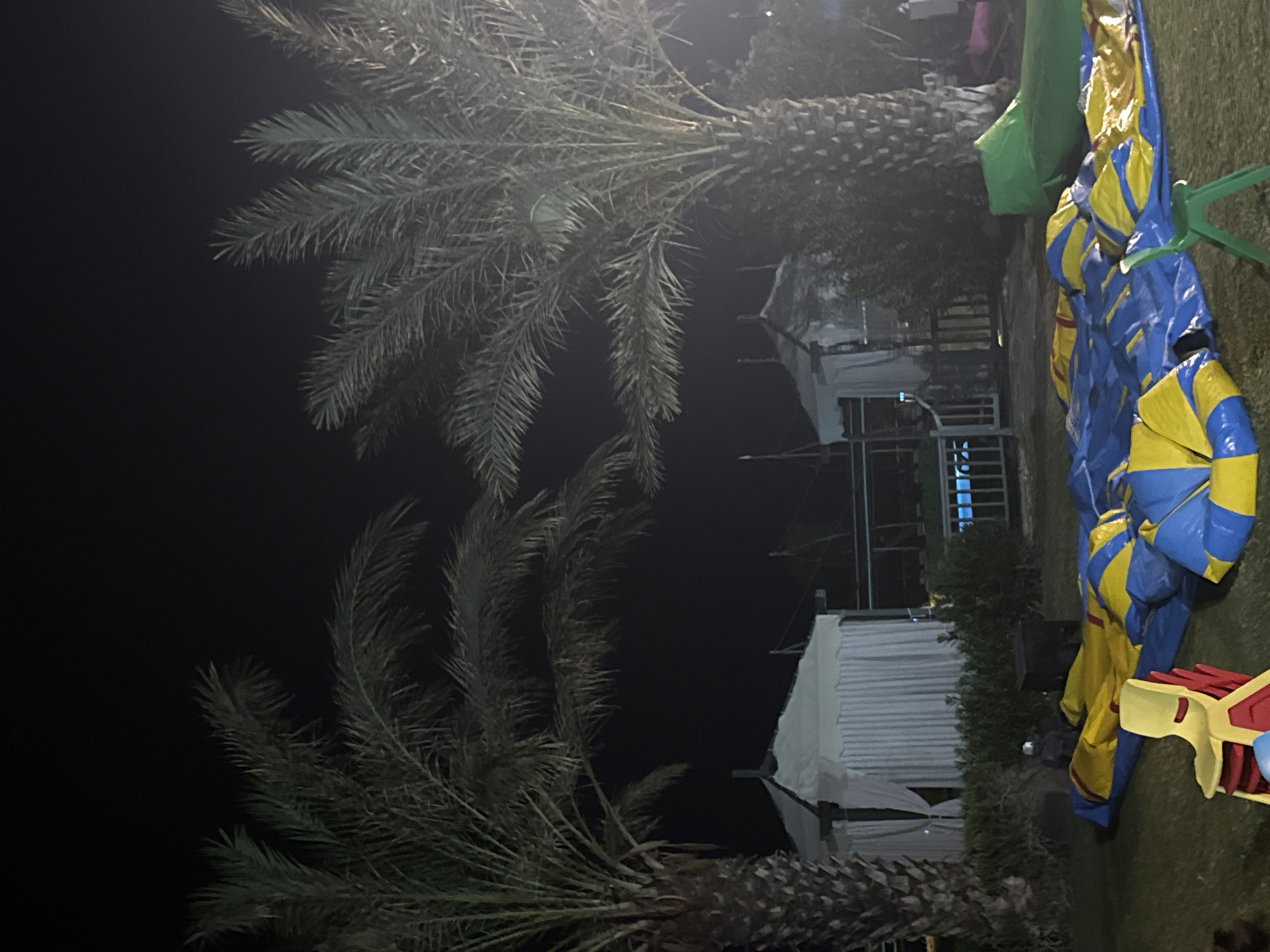 The image depicts an outdoor scene at night, possibly a recreational area or event space, located in Aksu, Turkey. Two large palm trees are prominent, with their fronds silhouetted against the dark sky. In the background, two white, tent-like structures with draped curtains are visible, suggesting temporary shelters or booths. A portion of a blue-tiled swimming pool is also discernible between the tents. In the foreground, a deflated inflatable structure, predominantly blue and yellow, lies on the grass. A stack of red and yellow children's chairs is positioned to the left of the inflatable. A green beanbag chair is situated to the right of the inflatable. The overall setting appears to be an outdoor venue prepared for an event, with some elements still being set up or taken down.