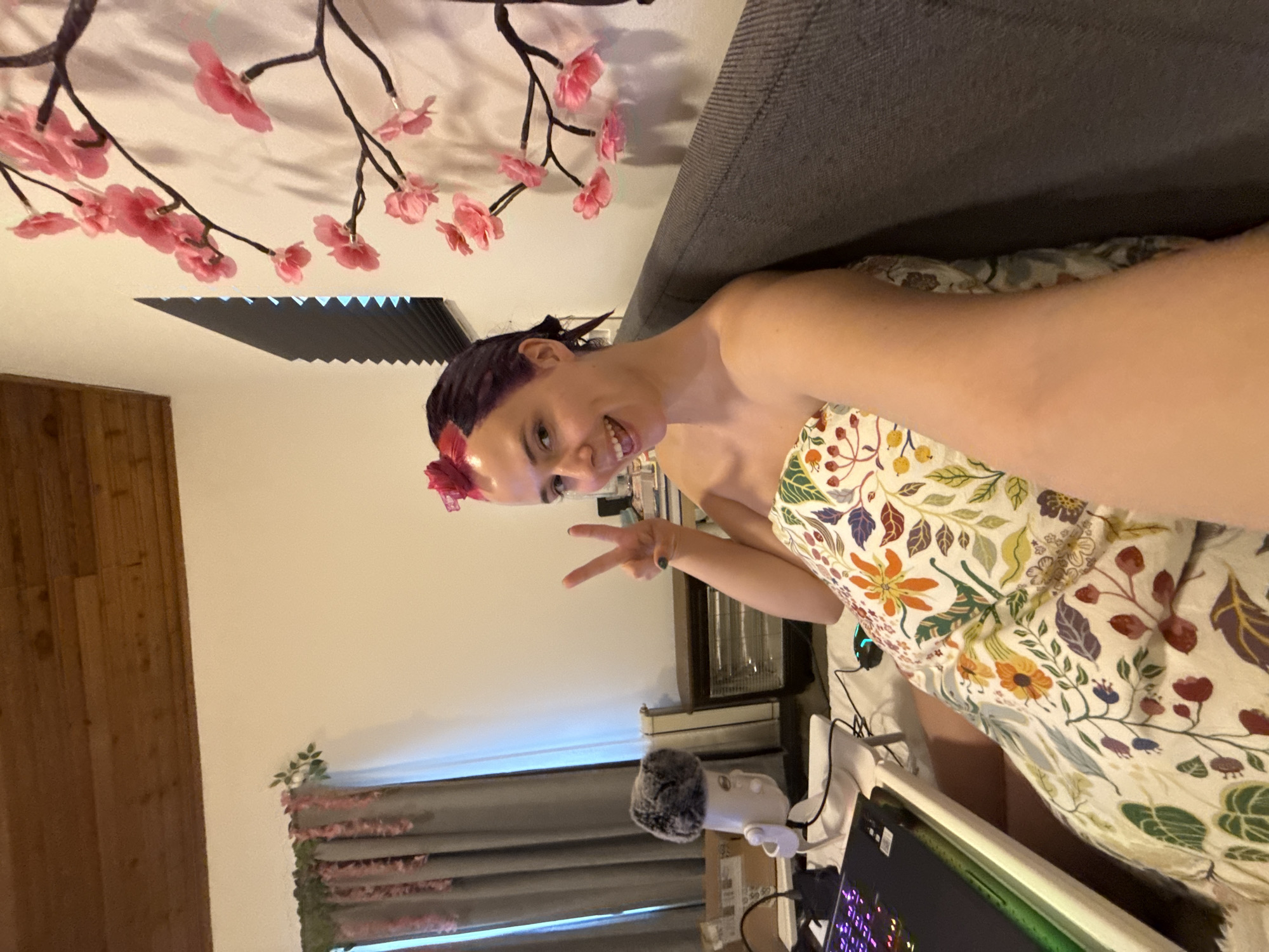 A woman is smiling and holding up a peace sign. She has purple hair and a flower in her hair. She is wearing a floral dress. She is sitting on a grey couch. There are faux cherry blossom branches on the wall behind her. A microphone, keyboard, and monitor are visible. There is wood paneling on the wall on the left. A window with blinds is visible. The image doesn't contain enough geographical context to determine the specific location in Zagreb, Croatia.
