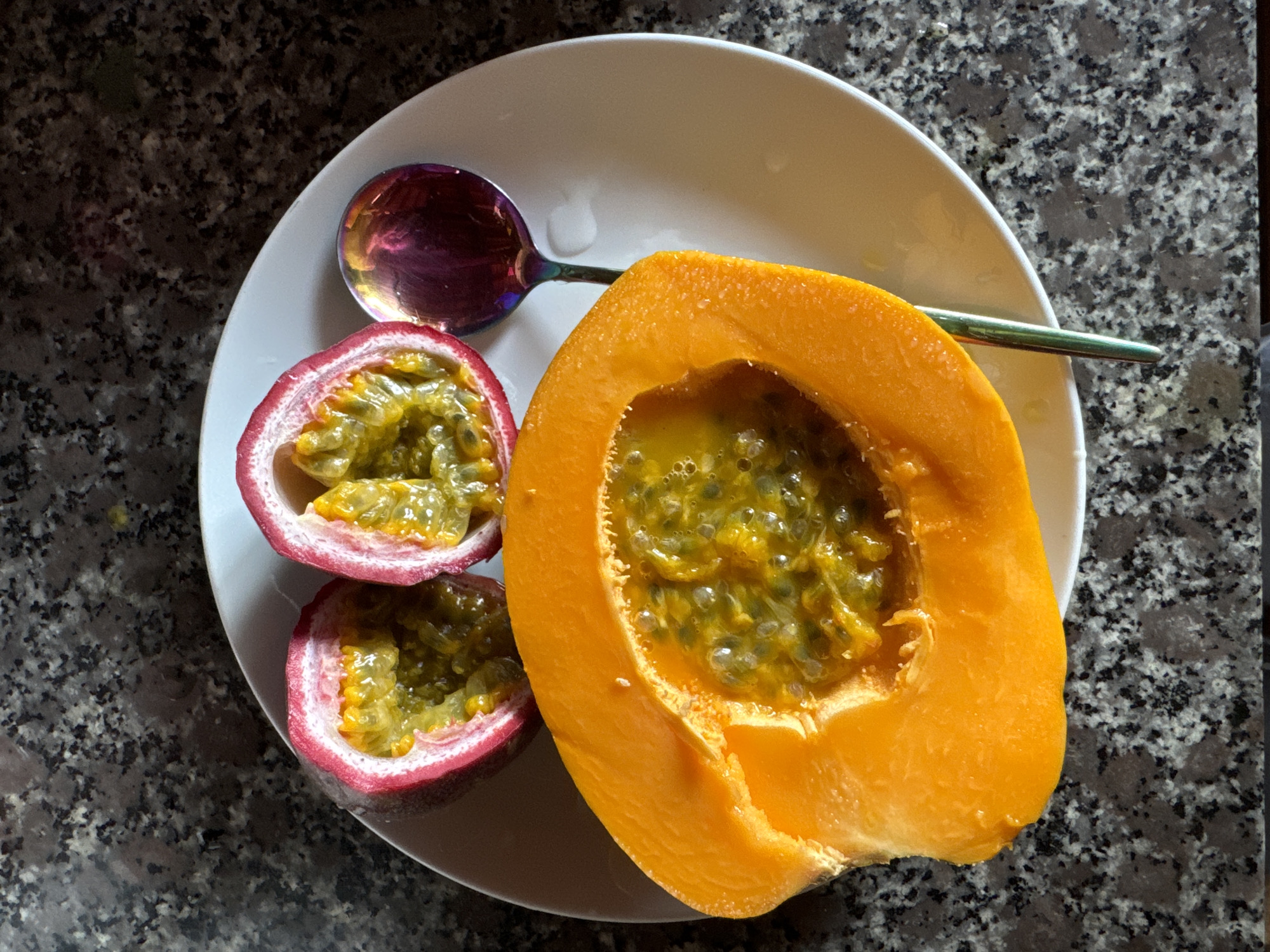 An overhead shot depicts a white plate containing two halves of a passion fruit, a halved papaya, and a spoon. The passion fruit halves have a reddish-purple rind and are filled with yellow pulp and black seeds. The papaya half has an orange rind and a similarly seeded pulp filling the cavity. A spoon with an iridescent bowl and green handle lies on the plate. The plate rests on a speckled gray and white surface, possibly a countertop. The location is assumed to be Mũi Né, Vietnam, based on the prompt's instructions, although there are no explicit indicators in the image. No people are visible.