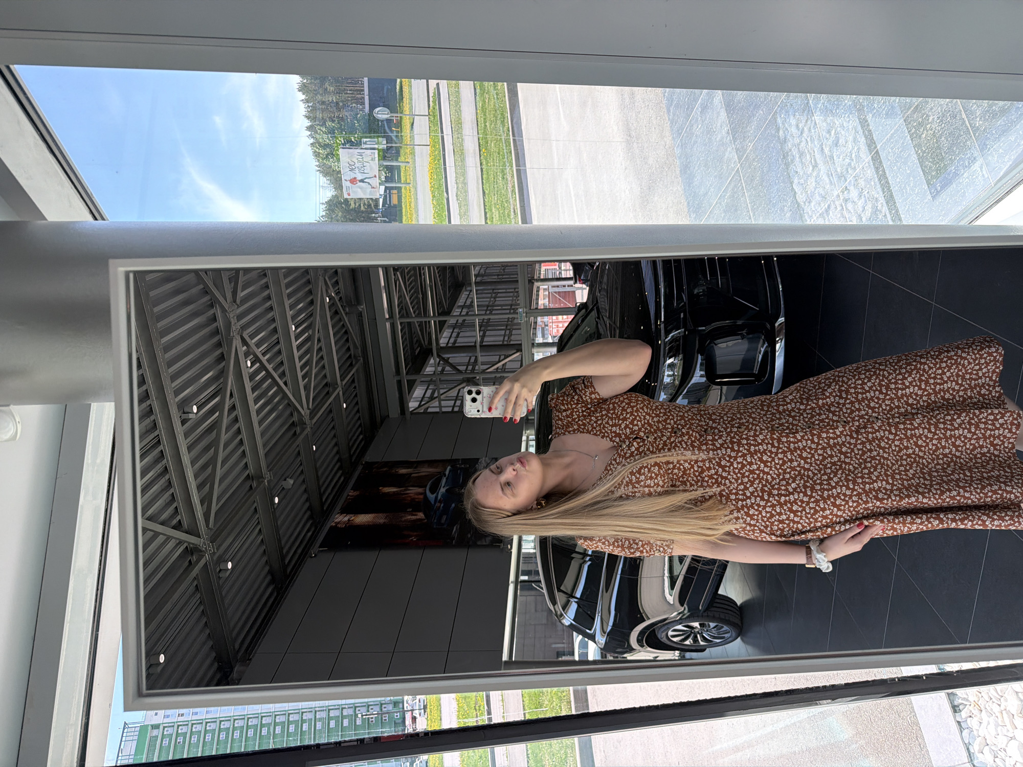 A woman is taking a selfie in a mirror inside a car dealership. The reflection in the mirror shows a black car and parts of the building's interior, including the ceiling structure and promotional posters. Through the window, a cityscape is visible, with signage reading "НСС Реклам." Based on the sign, the location is likely Izhevsk, Russia.