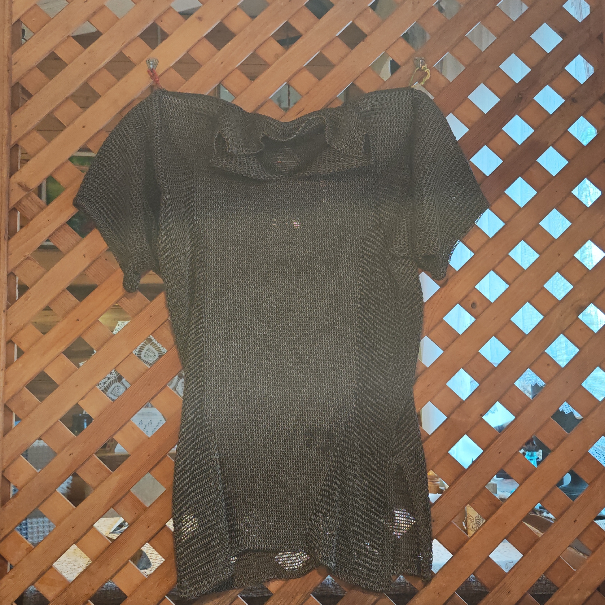 A short-sleeved, black chainmail shirt with a ruffled collar hangs on a wooden lattice. The lattice is mounted on a wall. The location appears to be in Rostov, Russia based on background objects seen through the lattice. The shirt is suspended by red and yellow cords from the top corners of the lattice. The shirt appears to be woven from small metal rings. Patches of light can be seen through the holes in the chainmail.