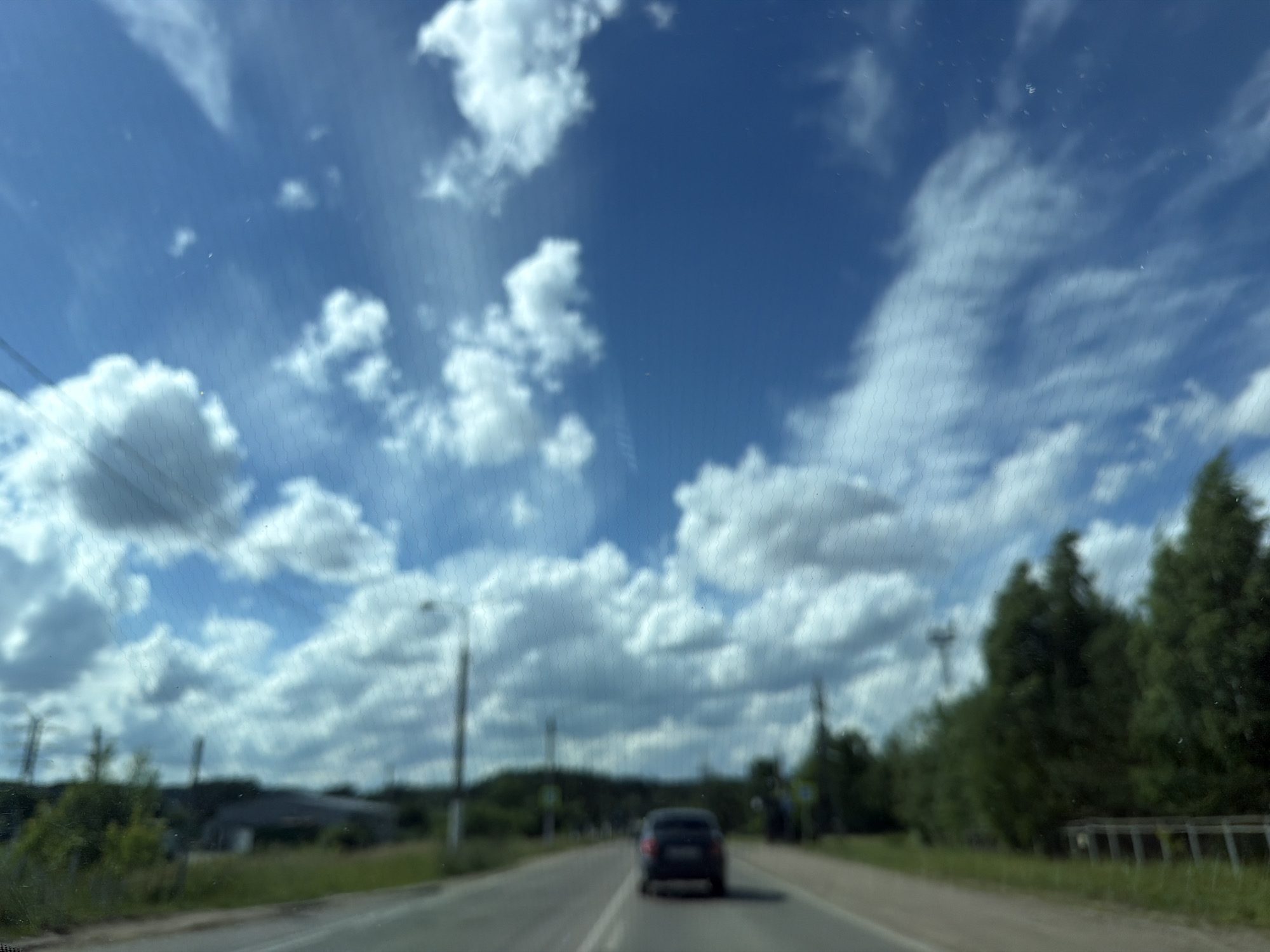 The image depicts a view from a vehicle traveling on a road, likely in the city of Chaikovsky, Russia, on a bright, cloudy day. The sky is a vibrant blue with scattered white cumulus clouds, some of which are wispy and streaky. In the foreground, the road stretches ahead, with visible lane markings. A dark-colored car is driving in the same direction, slightly ahead of the viewer.
On either side of the road, there is green grass and vegetation. On the left, there are some distant buildings and what appear to be utility poles or towers. On the right, there is a line of dense green trees. The overall impression is that of a rural or suburban landscape. The image is somewhat blurred, suggesting it was taken while the vehicle was in motion. The texture of a windshield with a visible pattern, possibly from dirt or rain, is also present in the foreground.