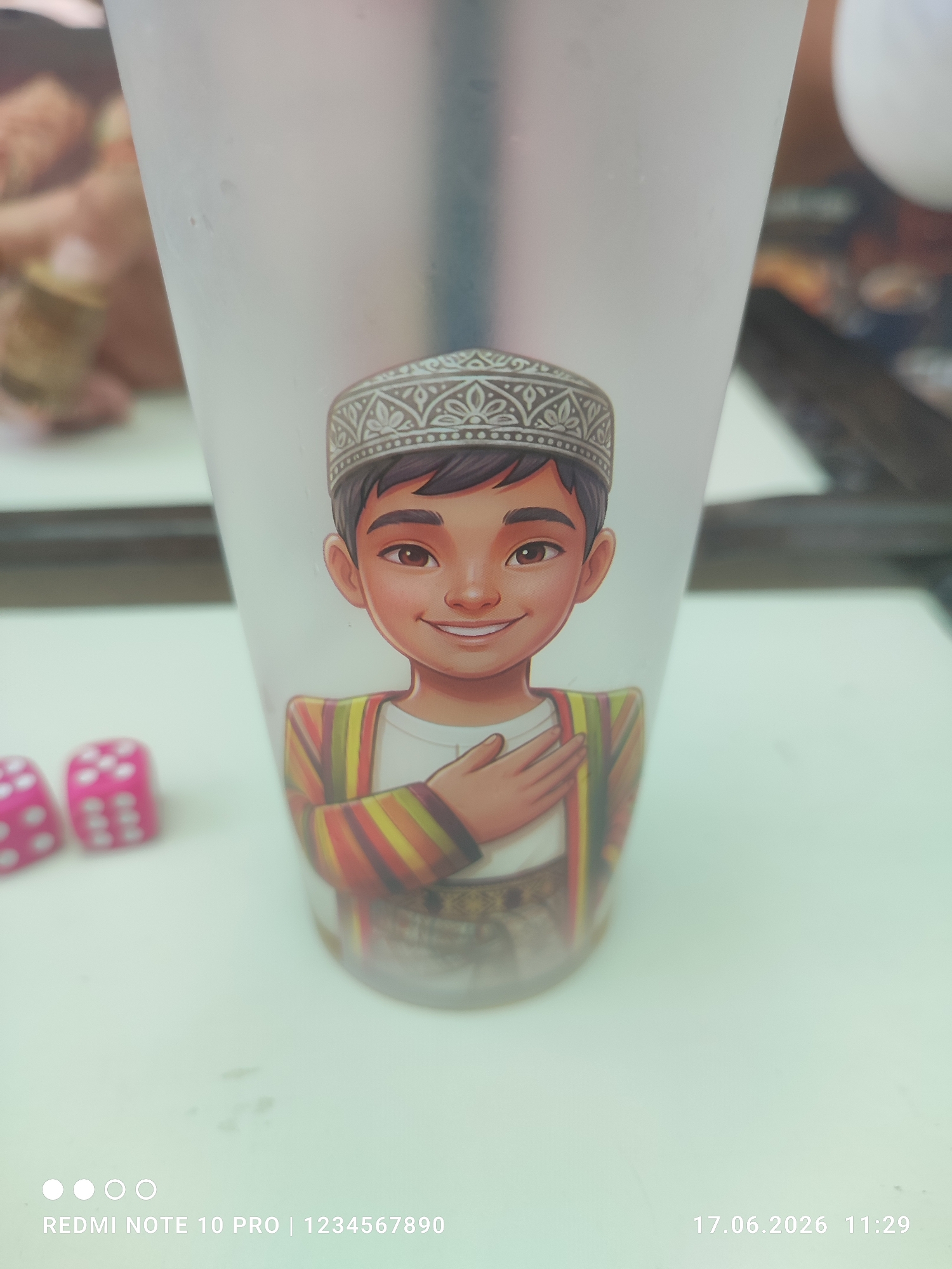 A cartoon of a young boy wearing a traditional hat and colorful striped robe is printed on a translucent cup. The boy has his right hand placed over his chest and is smiling. Two pink dice with white dots are visible in the lower left corner of the image, out of focus. The background is blurred and appears to be an indoor setting. The bottom of the image contains text and a date/time stamp.