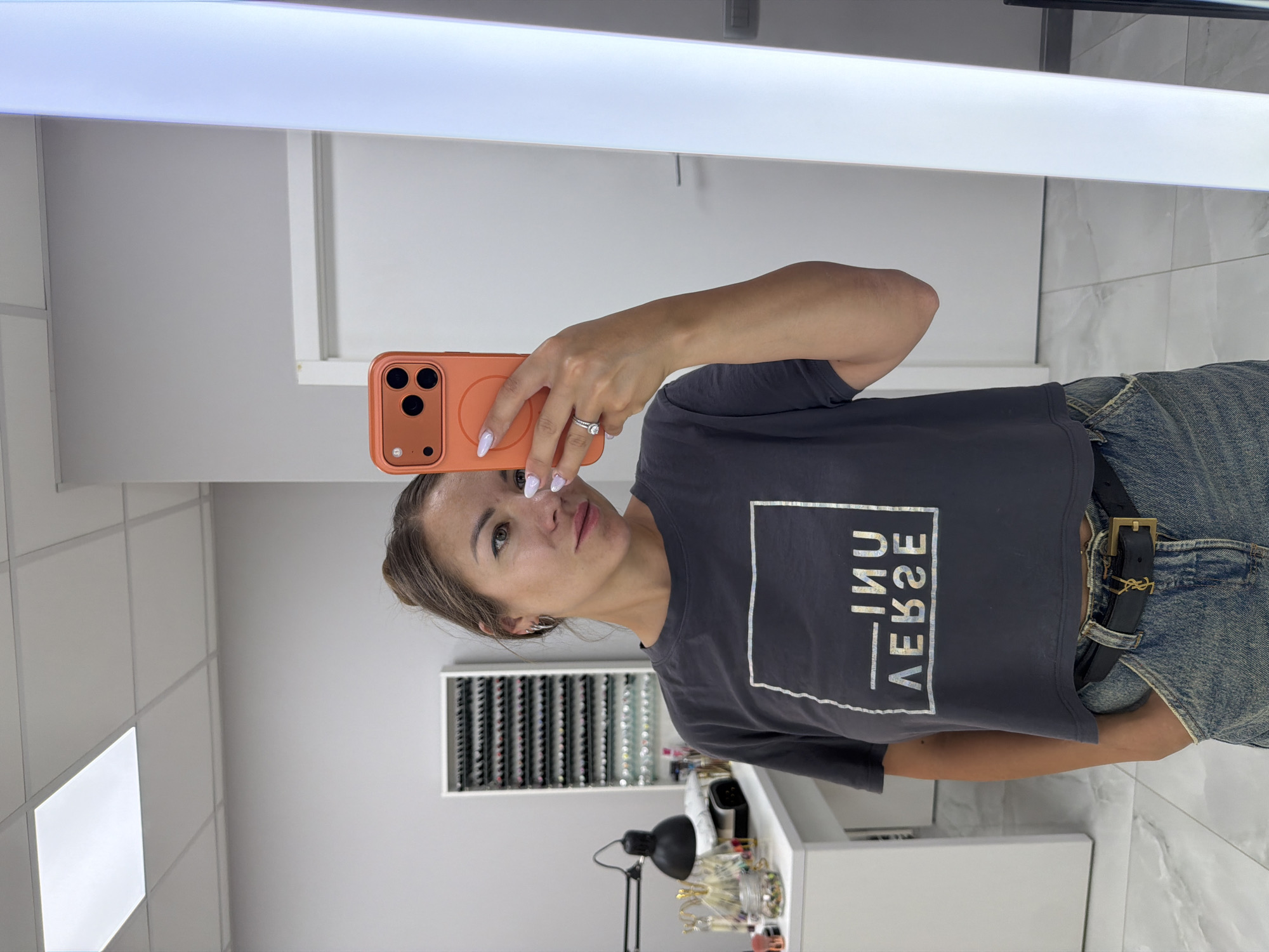 A woman with brown hair tied up in a bun, wearing a dark gray cropped t-shirt and blue jeans, takes a mirror selfie. She is wearing a black belt with a gold buckle and a ring on her left hand. She is holding an orange smartphone in her right hand, pointing it at the mirror. The background appears to be a brightly lit room with white walls and a white ceiling with fluorescent lighting. To her left, there is a white desk with a black lamp and various items on it, including jars and bottles. A tall, white shelving unit filled with small bottles is mounted on the wall behind the desk. To her right, a tall, bright vertical light fixture is visible. The floor is tiled with a marble-like pattern. The image is taken in what appears to be a salon or beauty studio. The scene is located in Budva, Montenegro.