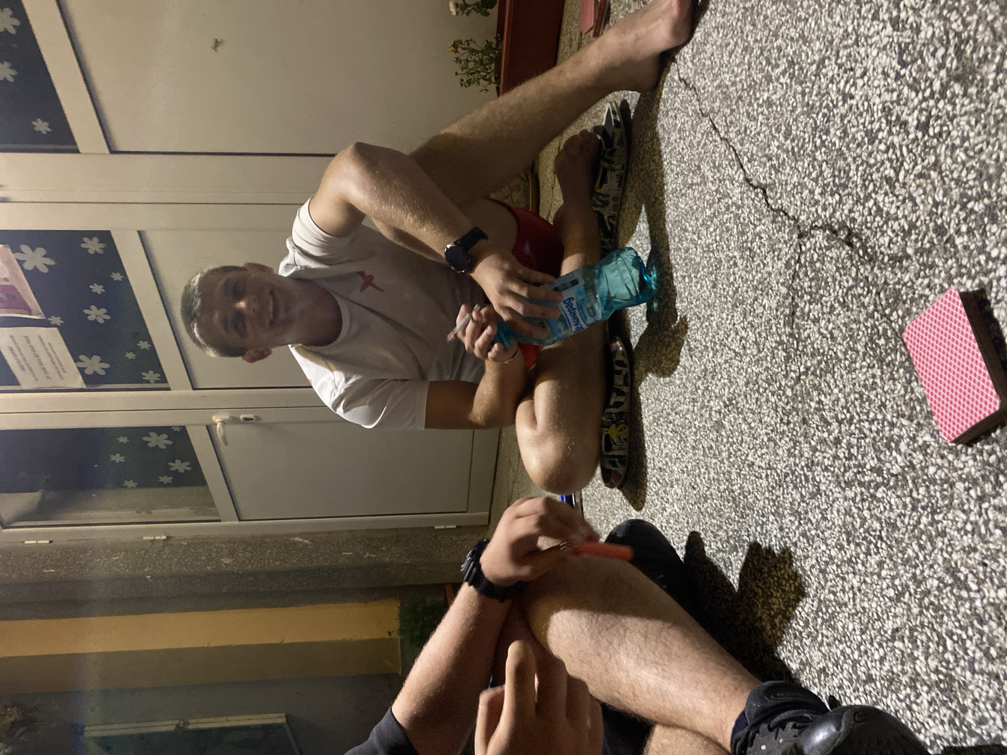 The image depicts a casual outdoor scene at night, likely in the city of Hirevo, Bulgaria, given the context provided. Two individuals are seated on the ground. One, a young man with short blonde hair, is smiling at the camera. He wears a white t-shirt and red shorts. Beside him, he holds a blue plastic water bottle and appears to be smoking. His right arm rests on his bent knee, and a watch is visible on his left wrist. The other person, whose body is partially visible on the left, is wearing black shoes and has a watch on their left wrist. This individual's hands are engaged with an orange object, possibly a pen or cigarette. A deck of cards with red backs is placed on the ground in the foreground. The background consists of a building entrance with white doors and windows decorated with white flower decals. The ground is paved with a textured material that appears to be concrete or asphalt with small stones. Plants in a brown planter are visible to the right. The lighting suggests it is evening or night, with artificial light illuminating the scene.