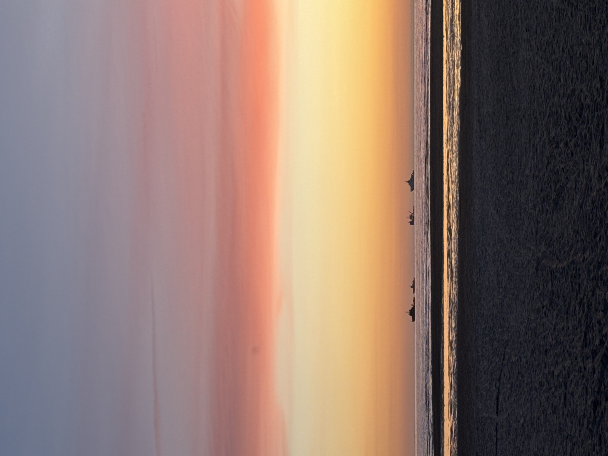 The image depicts a serene seascape at dusk, with the sky transitioning through shades of blue, pink, and orange. In the foreground, dark, wet sand marks the edge of a beach. Beyond the sand, a calm body of water stretches to the horizon, reflecting the warm hues of the sky. Several ships, silhouetted against the twilight, are visible in the distance. One cluster of ships appears to be a tugboat with a barge, while other vessels are more indistinct. The overall impression is one of tranquility and the peaceful end of a day. There are no people visible in the image. Based on the typical aesthetic of Baltic sunsets and coastal scenes in Estonia, this image could plausibly be from the coast of Tallinn, Estonia, perhaps near Pirita or along the Bay of Tallinn, where such unobstructed sea views are common.