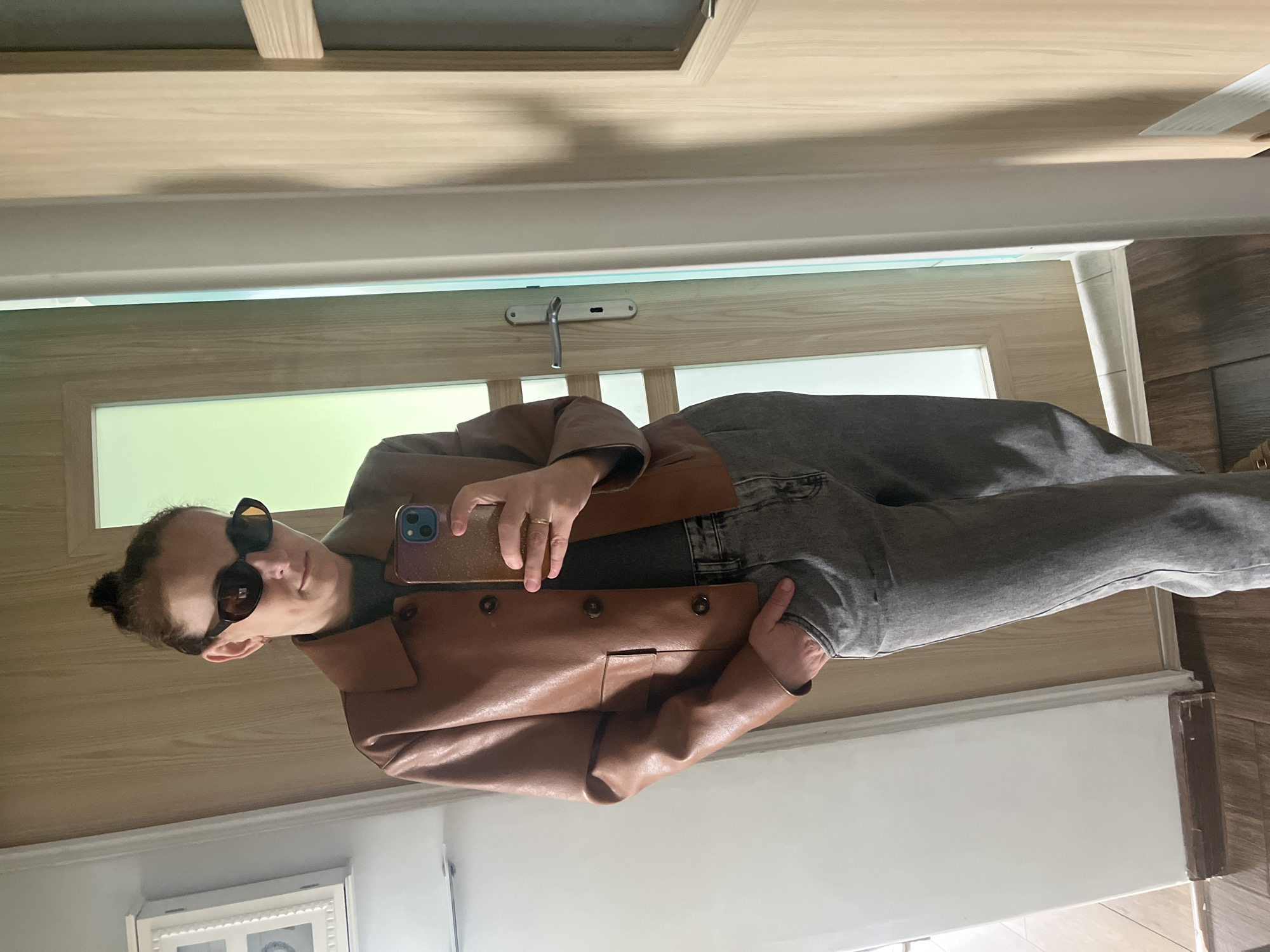 A person wearing sunglasses and a brown leather jacket stands in a doorway, taking a selfie with a smartphone. The person has their hair in a bun. The doorway is made of light wood and has a window near the top. The person is also wearing gray jeans. The location is Unknown, Unknown.