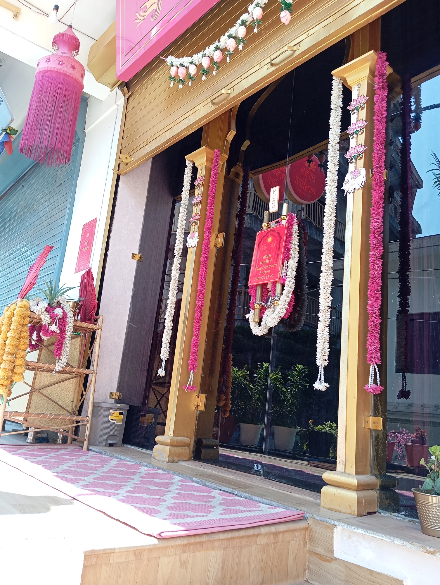 The image depicts the exterior of a business, likely a restaurant, located in Jonk, India. The building has a gold-colored facade with a closed roll-down door and glass windows. The entrance is decorated with garlands of flowers, both white and pink. A sign hanging in the center of the doorway reads "maa malini", and the words "Closed for lunch 2-3pm" are printed on the sign as well, followed by a phone number. A pink tassel-like decoration hangs from the ceiling to the left of the doorway. A bamboo shelf stands to the left of the entrance, decorated with more garlands and flowers. A pink and white patterned rug covers the walkway leading to the entrance. The glass windows reflect the surrounding buildings and vegetation.