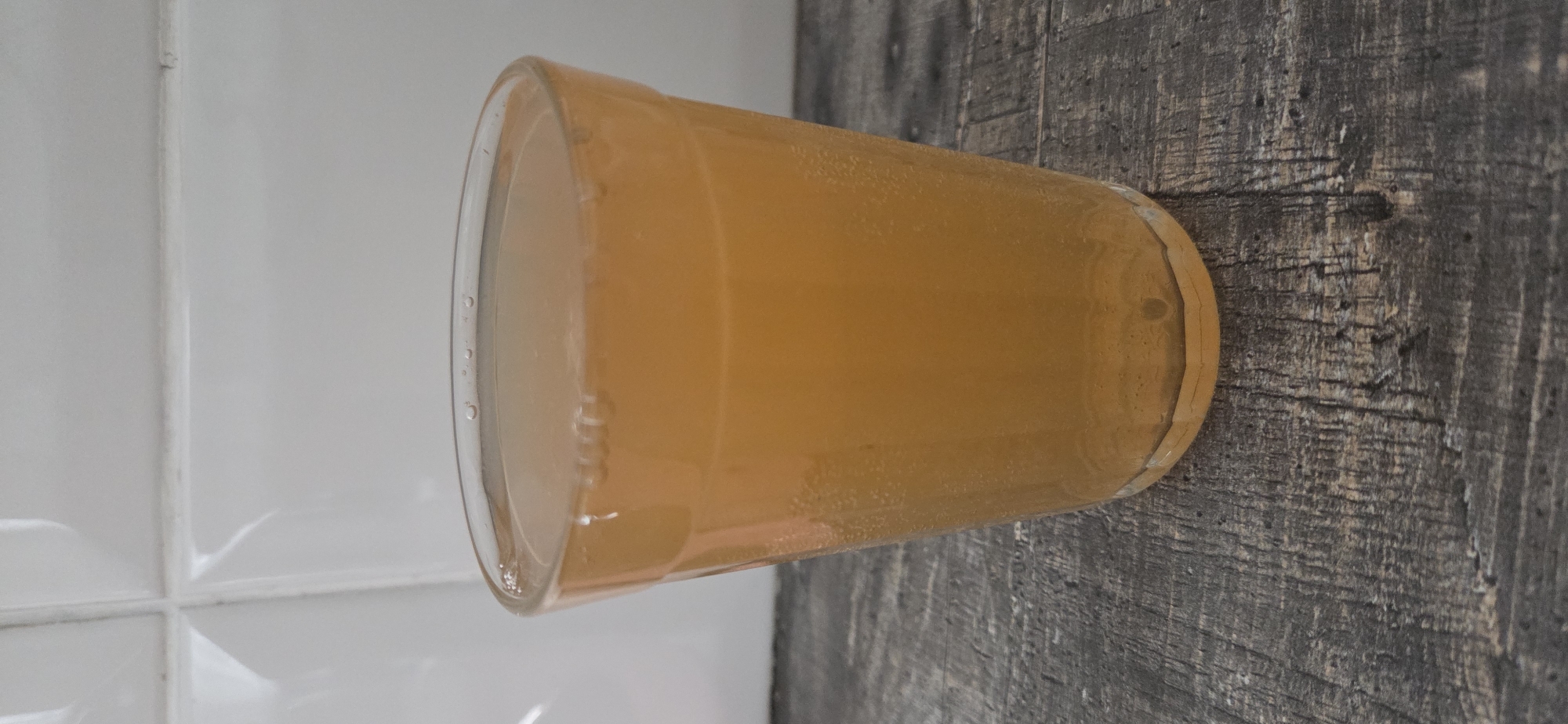 A clear glass containing a light brown, bubbly liquid is placed on a dark wood surface. The glass has a slightly flared rim and is filled to approximately three-quarters of its capacity. Small bubbles are visible both at the surface of the liquid and suspended within it. Behind the glass, a white tiled wall with dark grout lines is visible. The scene appears to be indoors, possibly in a kitchen or dining area. There is no indication of the specific location within the city of Lipetsk, Russia, as the image provides no landmarks or identifying features related to that locale. The image is a still photograph with no discernible action taking place.