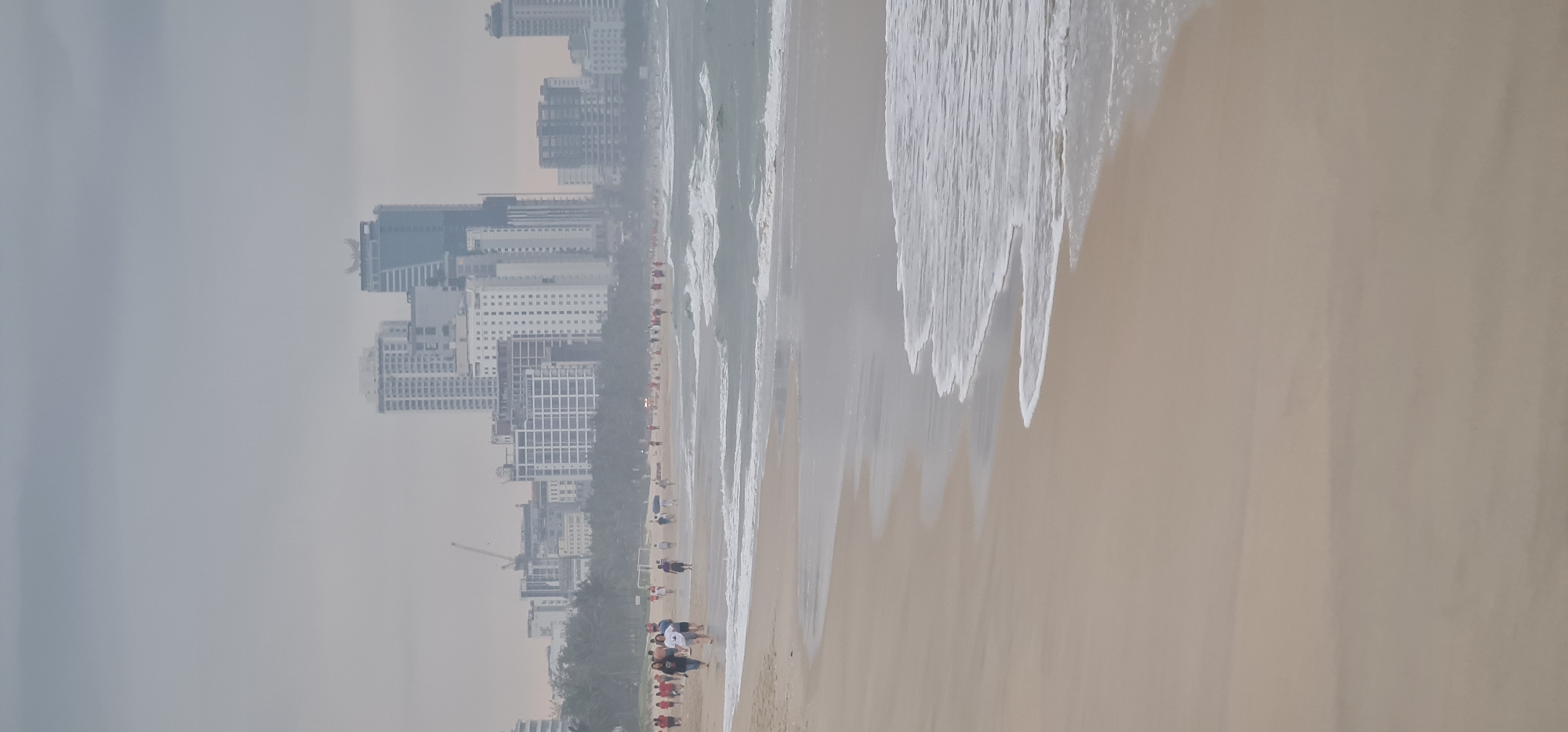The image shows a beach scene in Đà Nẵng, Vietnam, with a line of high-rise buildings in the background obscured by haze or fog. Waves are breaking on the sandy beach. People are seen along the shoreline. The sky is overcast, contributing to the overall hazy atmosphere.