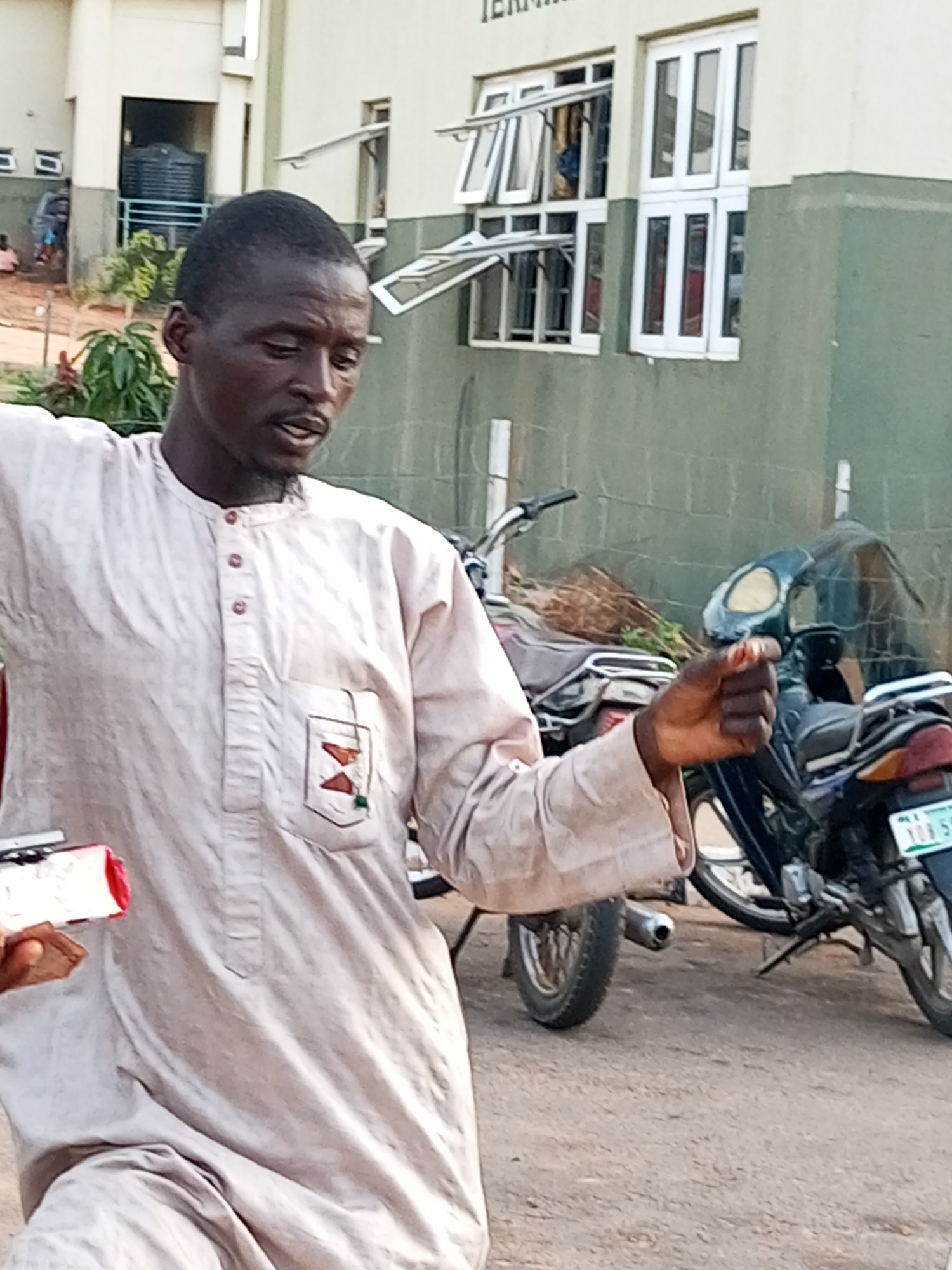 In Gombe, Nigeria, a man in a light-colored, traditional outfit stands outdoors, facing left with his head slightly tilted and his mouth open as if speaking or calling out. He has a dark complexion and a close-cropped hairstyle. His attire features a mandarin collar with three visible buttons and a square pocket on the left chest adorned with an embroidered emblem containing red and brown shapes. In his right hand, he holds a small, cylindrical object. Behind him, two motorcycles are parked on a dirt ground. The larger motorcycle on the right has a green license plate. In the background, a light-colored building with green accents is visible. The building has multiple windows, some of which are open, revealing interior spaces. The environment appears to be an outdoor urban setting.