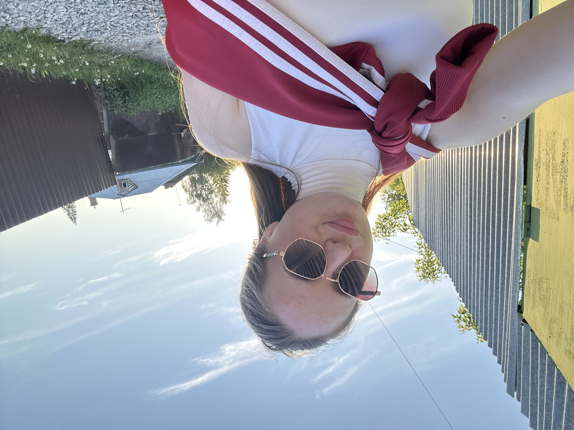 A young adult female, with long brown hair and visible freckles on her forehead, is depicted in a close-up selfie taken outdoors under a clear blue sky with scattered wispy white clouds. She is wearing octagonal-shaped sunglasses with rose-gold frames and dark lenses, and a white sleeveless turtleneck top. A red athletic jacket with three white stripes is tied around her shoulders and across her chest. Her lips are slightly puckered in a pout.
The scene is illuminated by strong, warm sunlight from the upper right, indicative of late afternoon or early morning. On the left side of the frame, a yellow-green painted vertical structure is partially visible, adjacent to a light grey corrugated metal fence that extends into the distance. Green foliage is visible behind and above this fence. The ground in the lower right foreground consists of a gravel path with patches of green grass and white dandelion puffballs. In the background on the right, a structure with a blue-grey corrugated metal roof and a small dormer window is visible, partially obscured by a darker, possibly corrugated metal fence. Distant trees, including deciduous and coniferous types, are present in the background. Utility lines are faintly visible against the sky in the upper right.