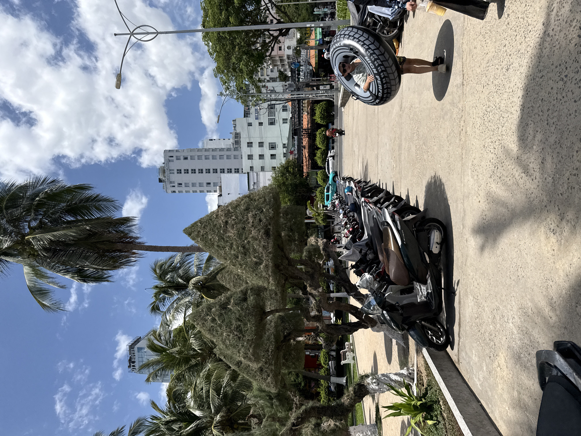 In Nha Trang, Vietnam, a young person is walking on a paved area, holding a large, black inflatable ring shaped like a tire. The ring has white lettering on it. The person is wearing a cap, sunglasses, and patterned shorts. They are walking amongst rows of parked motorcycles. In the background, there are tall buildings, including one with "OTX HOTEL" visible, palm trees, and a lamppost. The sky is blue with scattered white clouds. Another person is visible to the right, holding a drink. The scene appears to be a public space, possibly near a beach or resort area, given the presence of the inflatable ring and the tropical foliage.