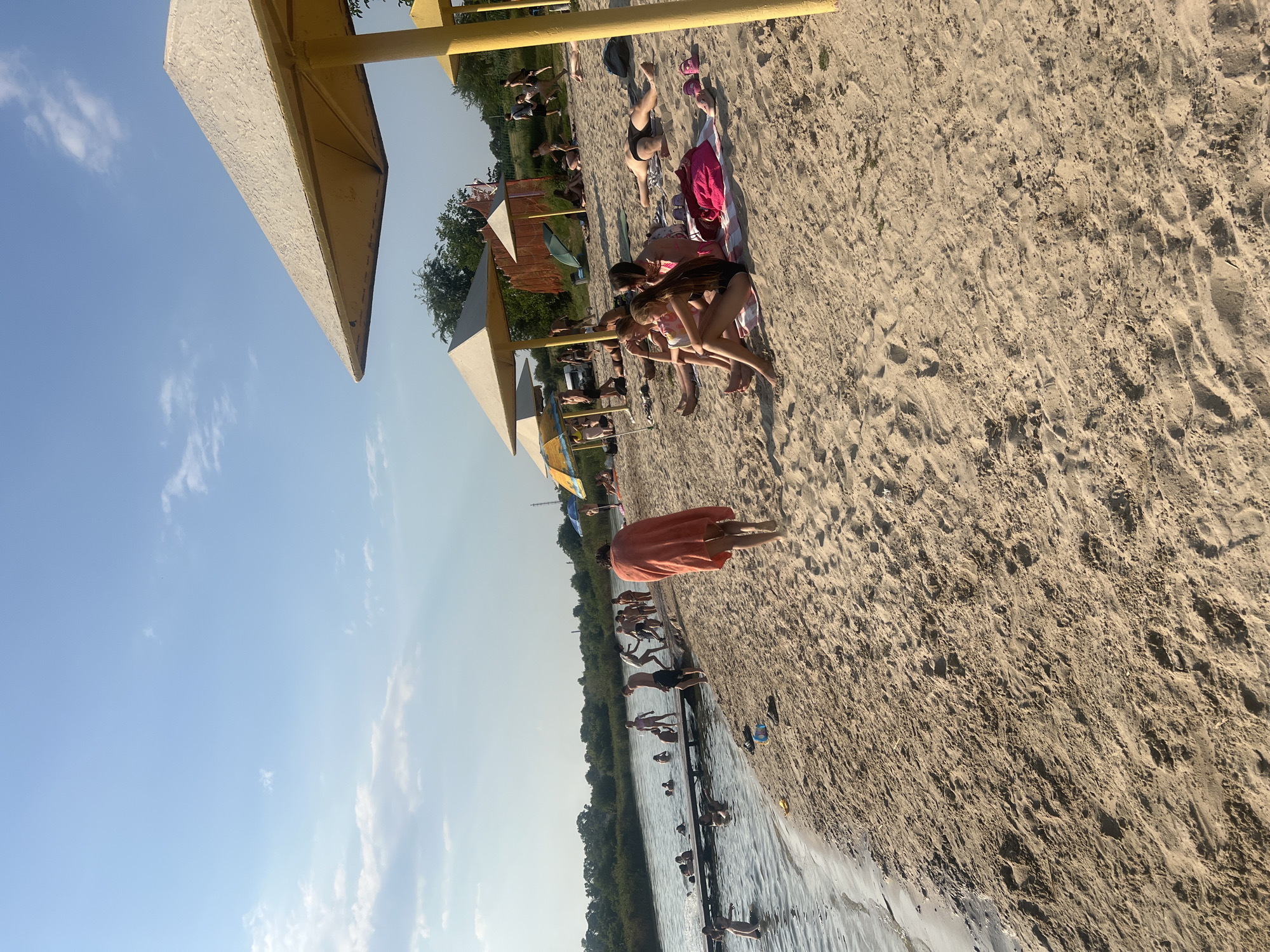 In an outdoor scene on a sandy beach, a group of people are enjoying a day by the water under a clear blue sky with scattered clouds. The beach is populated with individuals swimming in the calm water, walking along the shore, and relaxing on towels and under beach umbrellas. A wooden pier extends into the water, with several people standing on it or near it. The sand is textured with footprints and some small clumps of vegetation. In the foreground, a person wearing an orange towel walks away from the viewer. In the middle ground, several individuals are seated on the sand, some are talking to each other, and others are looking in different directions. Several beach umbrellas, some yellow and some with blue, provide shade. In the background, trees and foliage line the opposite side of the water, and more people can be seen in the distance, some playing near the water's edge.