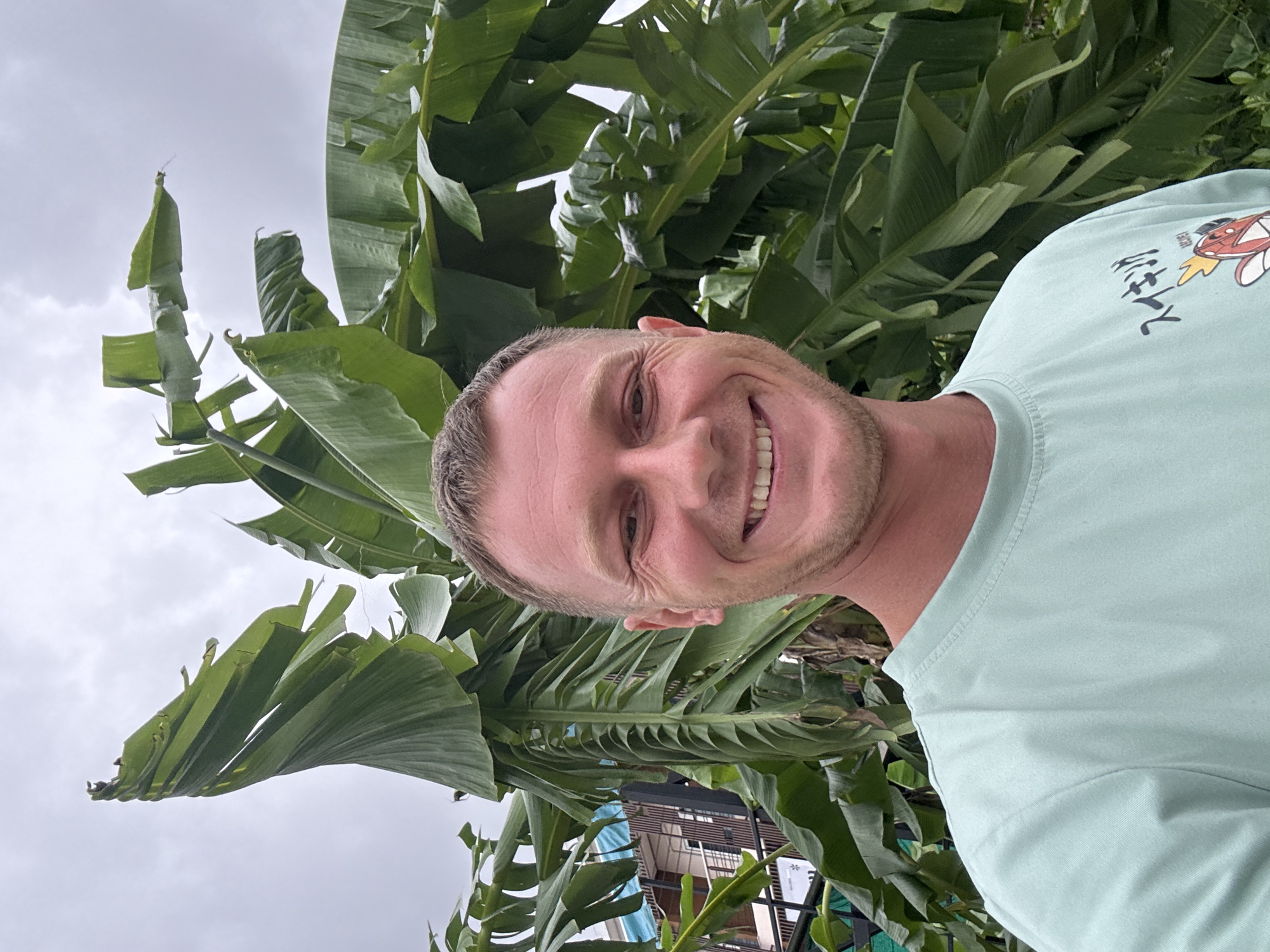 A close-up, selfie-style photograph features a fair-skinned man with short, blonde hair and stubble, smiling widely. He is wearing a light teal t-shirt with Japanese characters and a cartoon image on the left chest. The man is positioned in front of a backdrop of large, lush green banana leaves. In the background, partially obscured by the foliage, is a building with a light-colored facade and dark trim, appearing to be a multi-story structure, likely within a residential or commercial area. The sky above is overcast and grey. The overall scene suggests an outdoor setting in a tropical climate, consistent with locations like Chalong, Thailand.