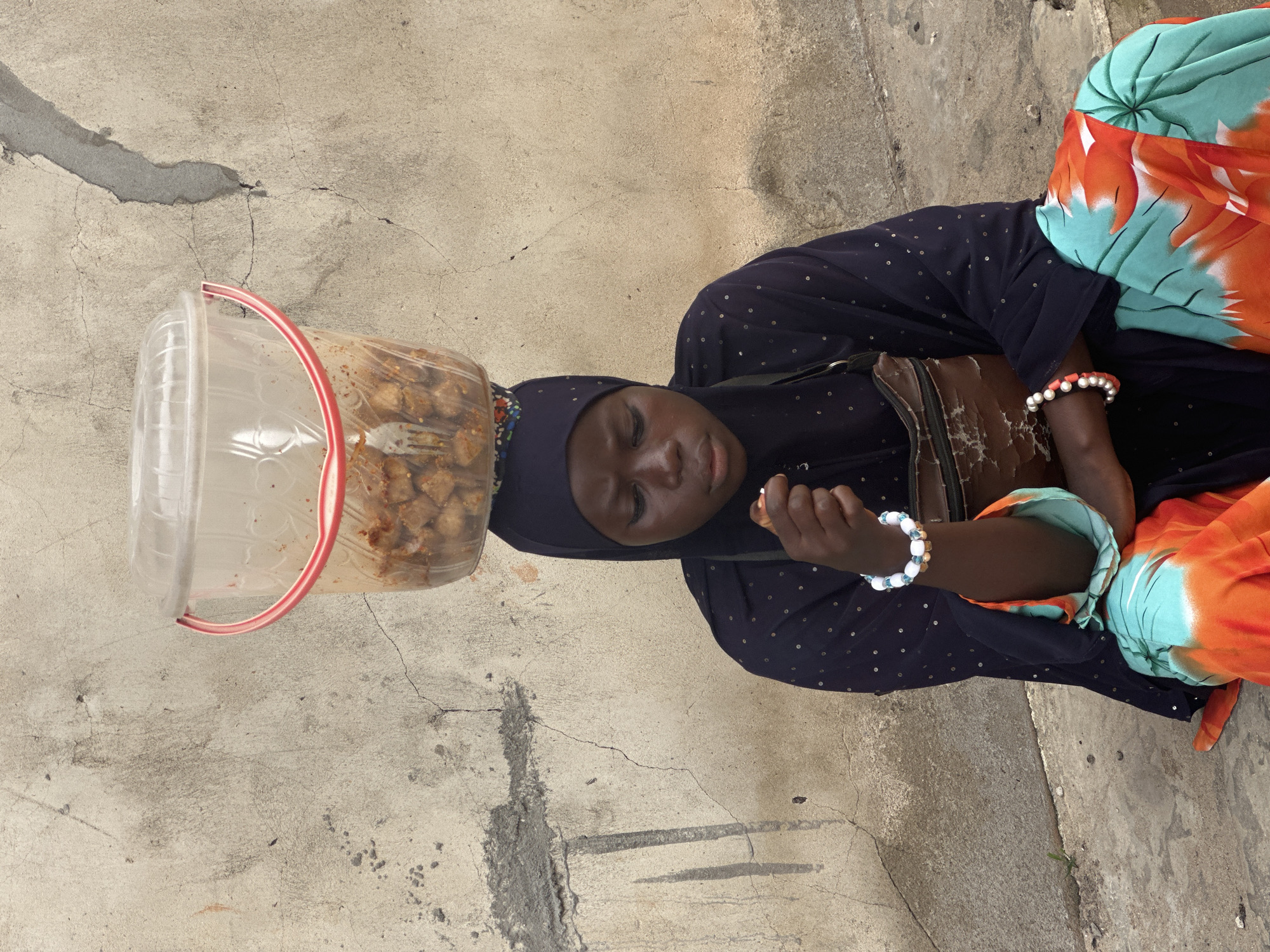 In Badagry, Nigeria, a young Black girl with dark skin and dark eyes, wearing a dark blue hijab, a dark blue garment adorned with tiny silver sequins, and a brightly colored orange and turquoise wrap skirt, sits on a rough concrete surface. Her head is crowned with a clear plastic bucket containing what appears to be food. She holds a small fork or skewer in her right hand, bringing it to her mouth as if to eat. On her left wrist, she wears a bracelet of white and blue beads, and on her right wrist, a bracelet of red and white beads. A worn brown leather pouch with a zipper is slung across her chest. The background is a textured, weathered concrete wall with visible cracks and a rough patch of grey cement. A small green sprout emerges from a crack in the ground to her left.