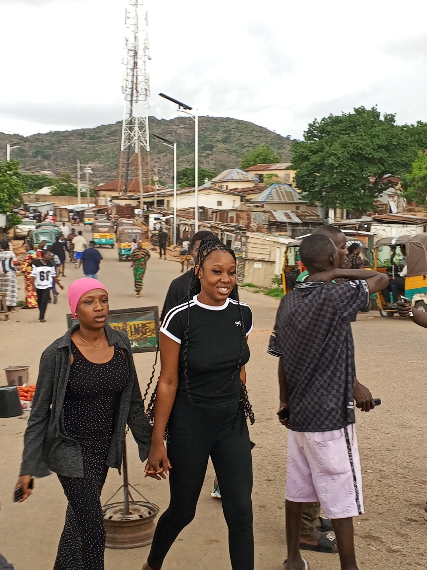 The image depicts a street scene in Gwong, Nigeria. In the foreground, two women are walking and holding hands. The woman on the left wears a pink headscarf, a black and white polka-dot dress, and a gray jacket. She holds a cell phone in her left hand. The woman on the right wears a black athletic outfit with white trim. Behind them is a man in a checker-patterned black and gray shirt and pink shorts with a black stripe. To the left of the main subjects, people walk along the street; several appear to be wearing traditional clothing. Yellow auto rickshaws are visible in the background. Buildings of varying heights are visible, and a mountain range is in the distance. A tall communications tower is visible above the buildings and a streetlight.