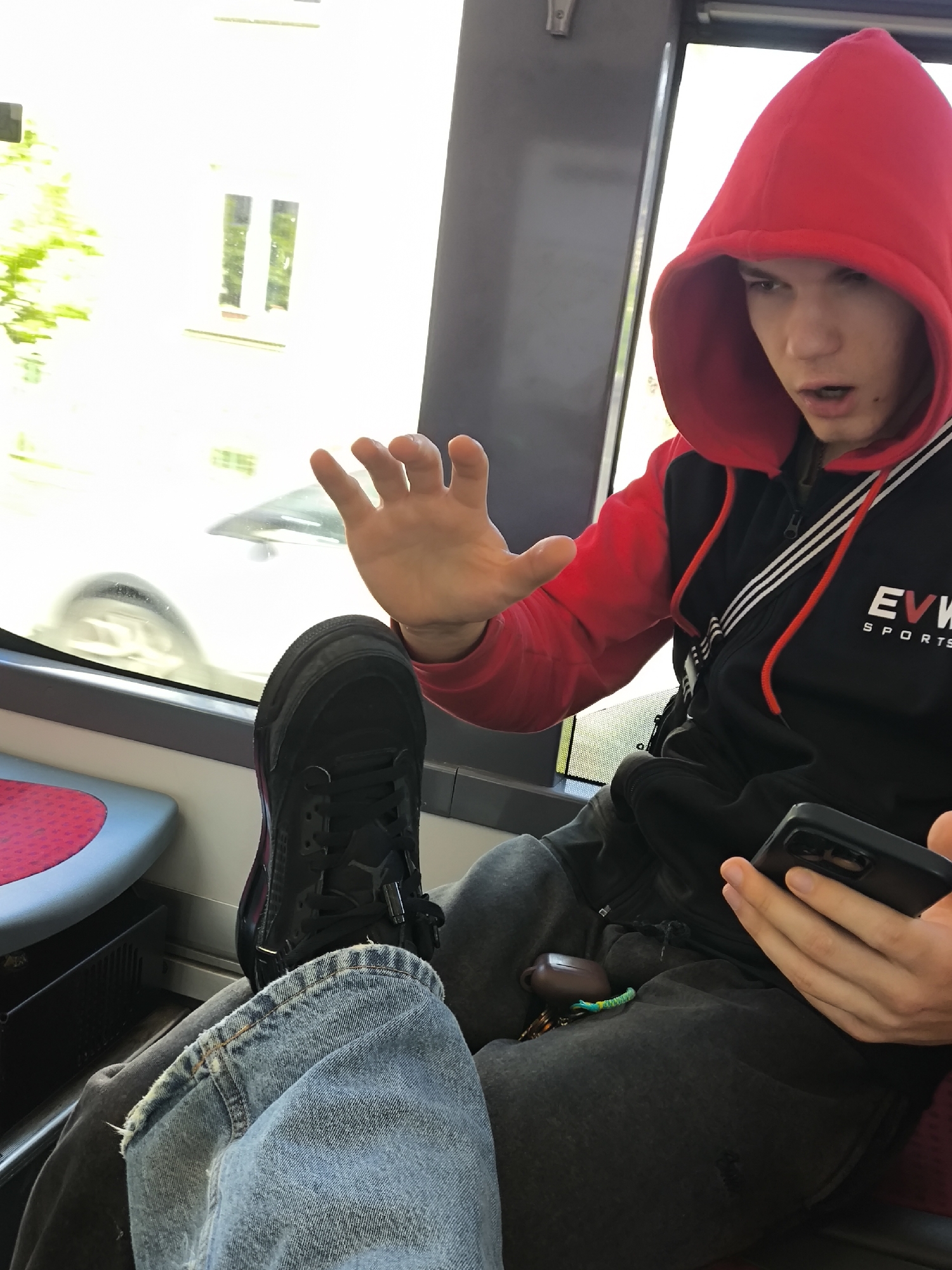 A young person, likely male, is captured in a candid shot inside a public transport vehicle, possibly a tram or bus, given the distinctive red-patterned seat and large windows. He wears a black zip-up jacket with white stripes on the shoulder and red drawstrings, topped with a bright red hood over his head. His expression is one of surprise or intense reaction, with his mouth slightly open and eyes wide, seemingly reacting to something out of frame.
His right hand is raised, palm open, in a gesture that suggests surprise, a halt, or emphasis during a conversation. In his left hand, he holds a black smartphone. His right foot, wearing a black sneaker, is propped up, revealing light blue ripped jeans on that leg, while his left leg is clad in dark grey sweatpants. On his lap, a brown case (likely for wireless earbuds) and keys with a green accessory are visible.
Through the window, a bright, sunny daytime scene unfolds, featuring blurred green foliage, a building with rectangular windows, and the indistinct form of another vehicle's wheel, suggesting motion. Visible text on the black jacket reads "EVV SPORTS". The interior of the vehicle includes grey wall panels. The setting is consistent with urban public transport in Strasbourg, France.