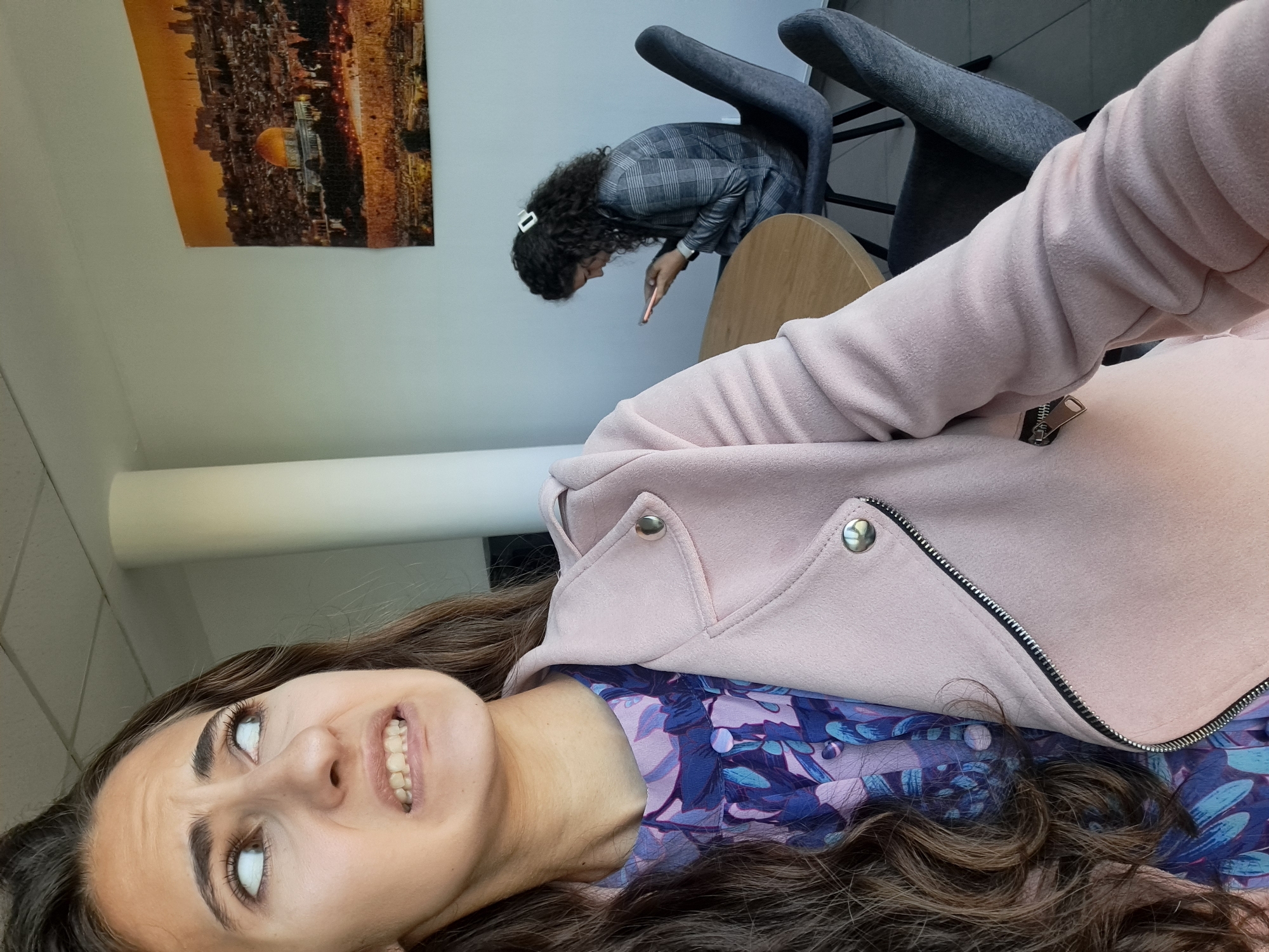 The image shows two young women in an indoor setting, likely an office or cafe, with a large framed picture of Jerusalem on the wall. The woman in the foreground is looking up and to the left with a distressed or disgusted expression, her teeth bared. She is wearing a light pink jacket over a blue and purple floral print dress. Her hair is dark brown and curly. In the background, a second woman with curly dark hair, wearing a grey checkered blazer, is seated at a round wooden table, looking down at her smartphone. The environment appears bright with natural light coming from the left. There are chairs and a table visible, suggesting a social or work-related space. The picture on the wall depicts the Dome of the Rock and the city of Jerusalem. There is no indication of Sofia, Bulgaria in the image.