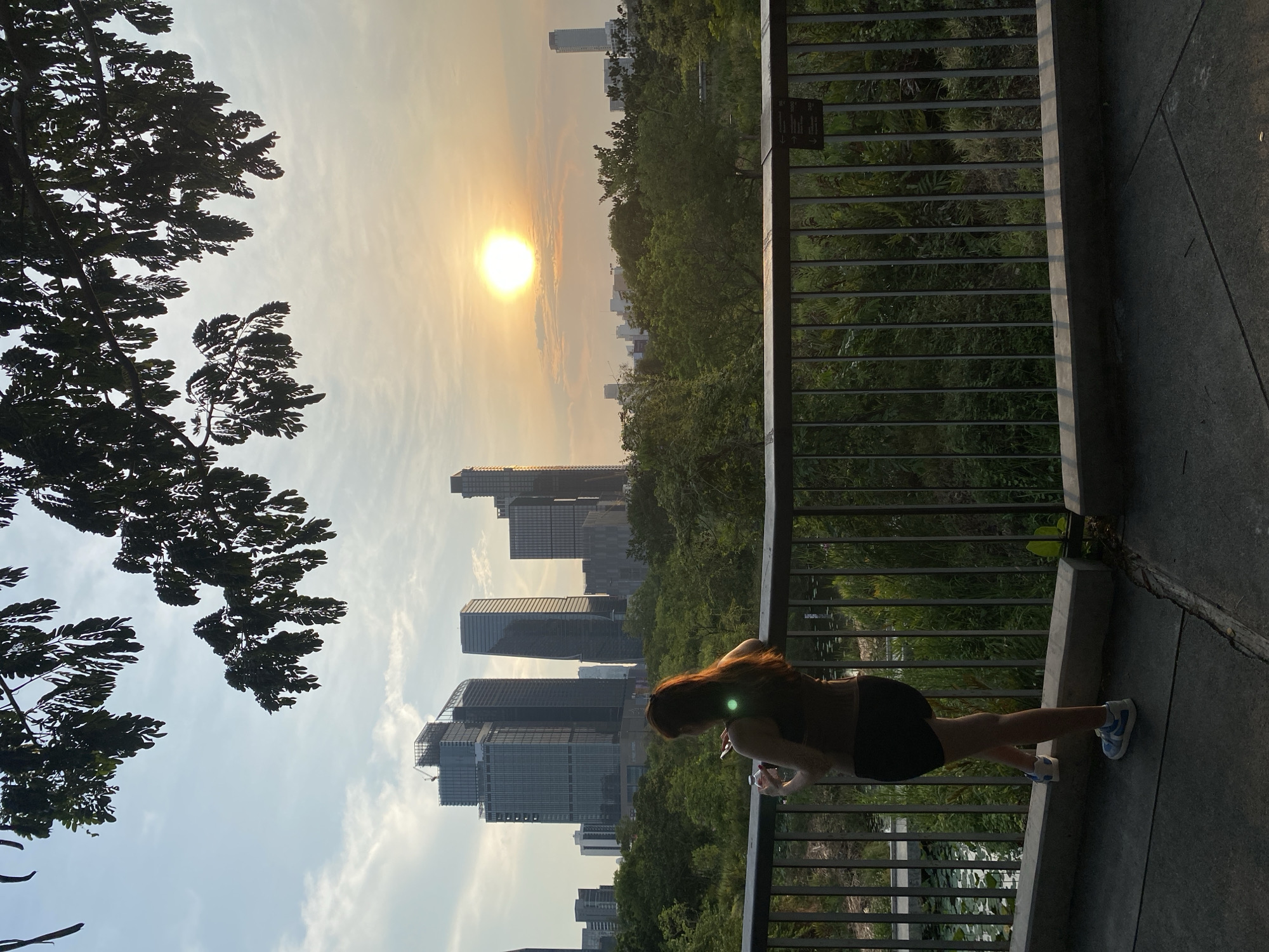 A slightly high-angle shot presents a cityscape at sunset, potentially in Bangkok, Thailand, as the building architecture resembles common Bangkok structures. In the foreground, a woman is seen holding onto a metal railing along a walkway. She has reddish-brown hair and is wearing dark shorts and a top. Her body appears to be facing away from the camera. The metal railing runs along the edge of a concrete walkway. Behind the railing, there is a dense green area with trees and foliage. In the background, several tall buildings are partially visible through the trees and near the setting sun. The sky is bright with the sun setting, casting a warm glow over the scene. A tree is on the left side of the image.